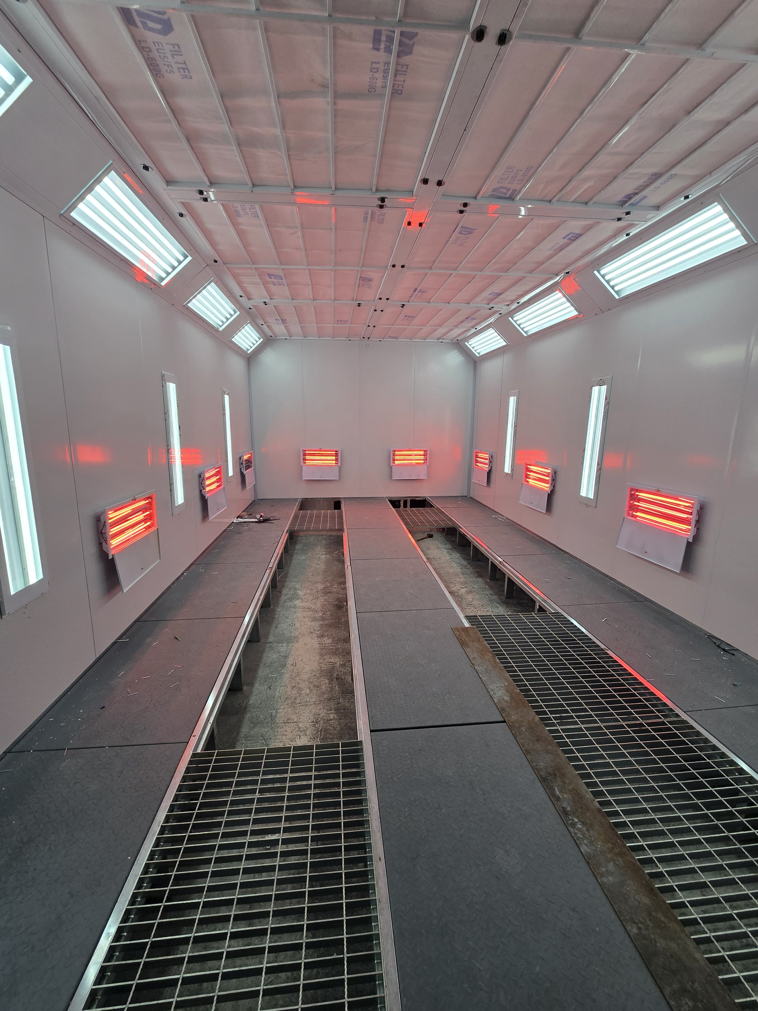 Innenansicht eines Labors oder einer Lackierkabine mit beheizten Infrarotstrahlern an den Wänden und Decke, Metallgitterböden sowie leere Arbeitstische.