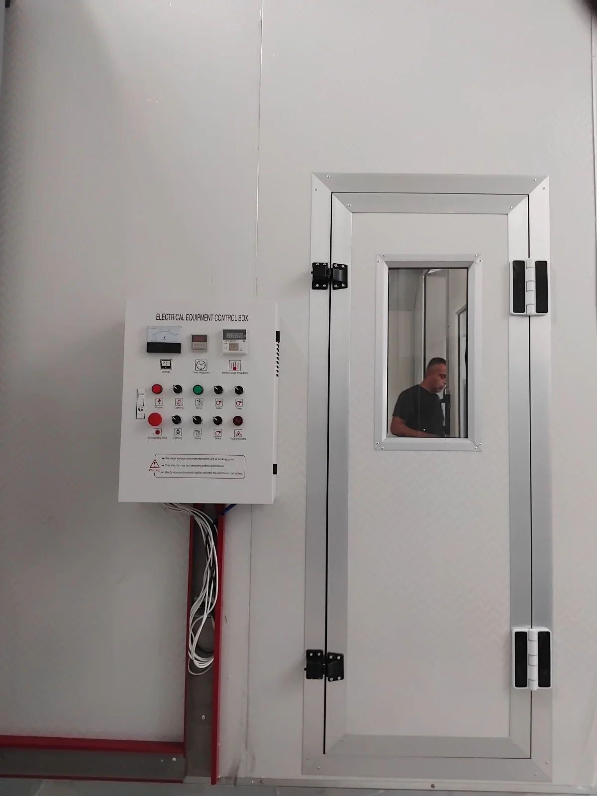 Ein weißer Kontrollkasten mit verschiedenen Schaltern und Anzeigen befindet sich an einer weißen Wand, neben einer großen weißen Tür mit kleinem Fenster und schwarzem Schloss. Im Fenster ist eine Person zu sehen.