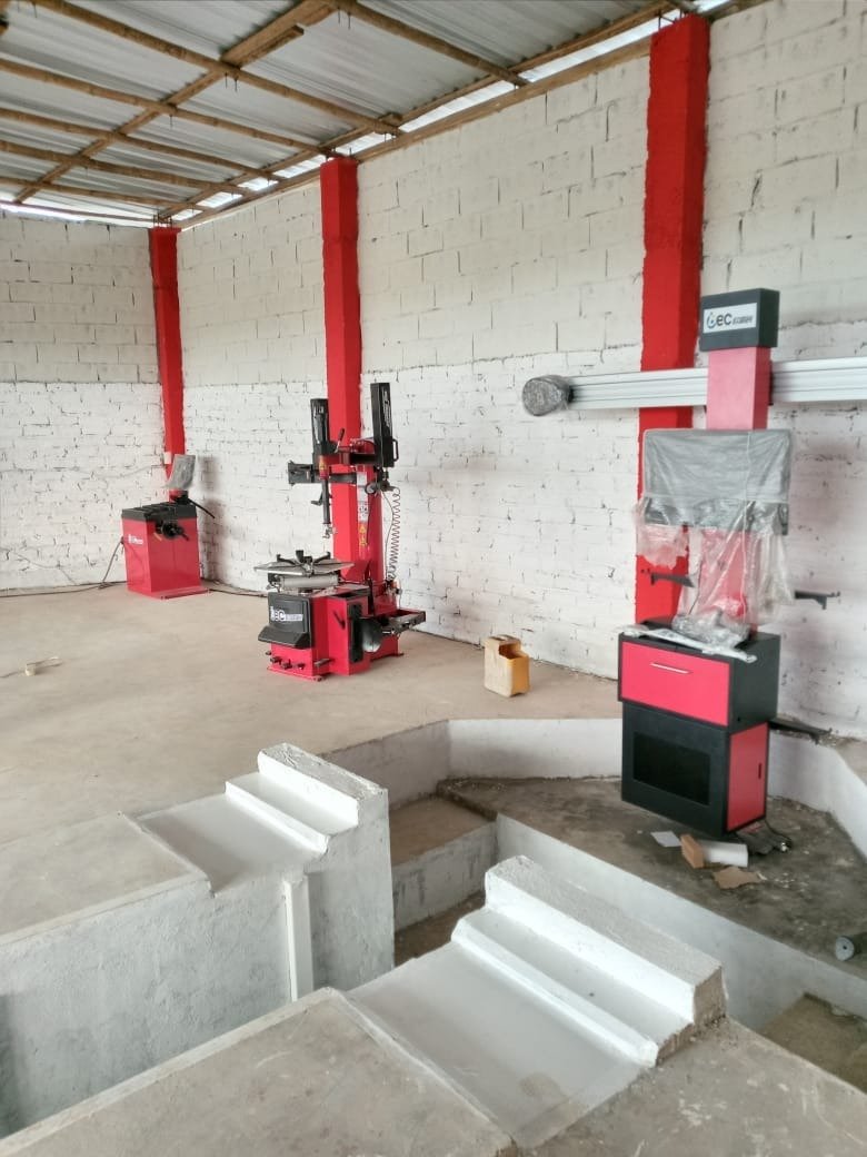 Werkstatt mit Rot-Schwarz Maschinen, weißen Ziegelmauern und Holzkonstruktion am Dach. Außerdem sind Betonschritte und eine Aussparung im Boden sichtbar.