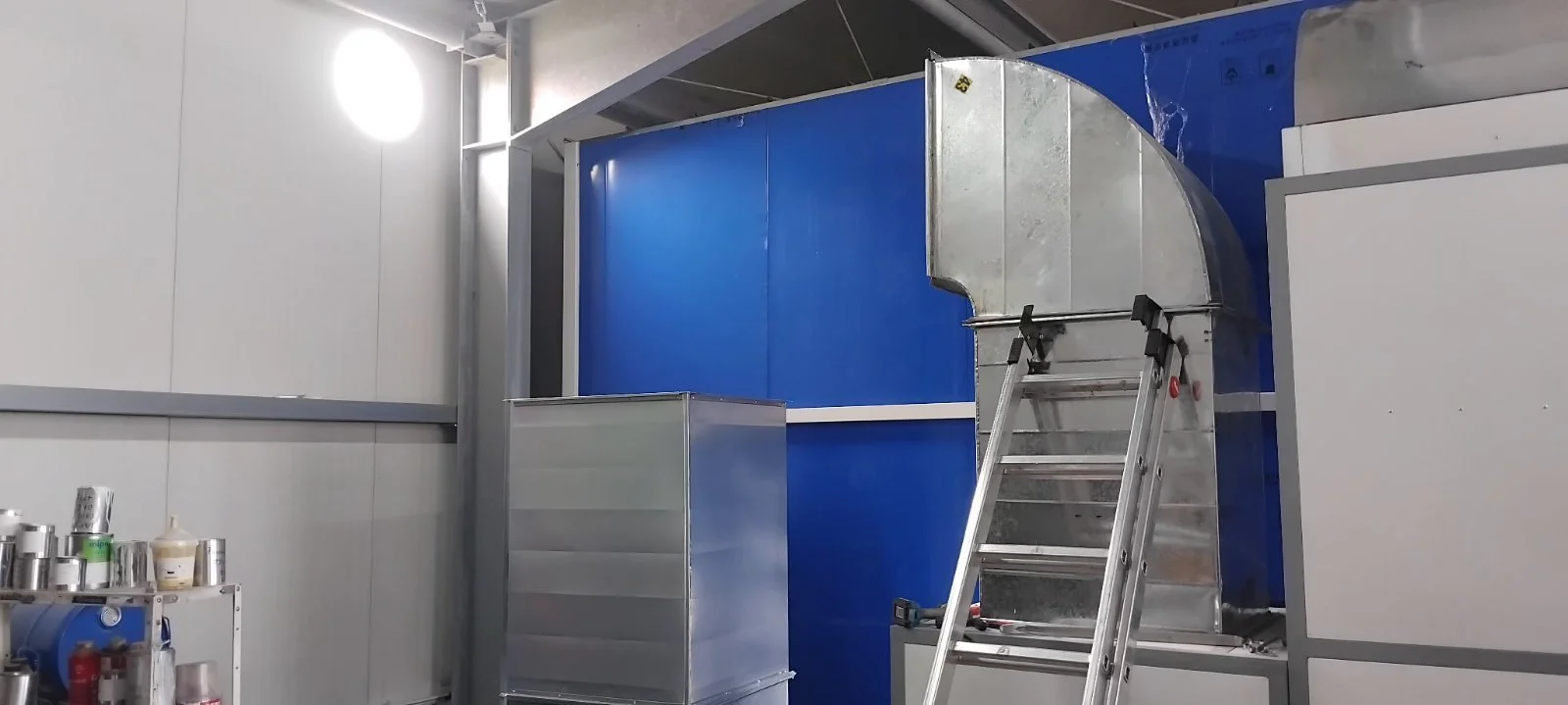 Innenraum eines Industrie- oder Lagerraums mit einer Leiter, einem blauen und silbernen Schrank, sowie Metallregalen mit Behältern und Flaschen.