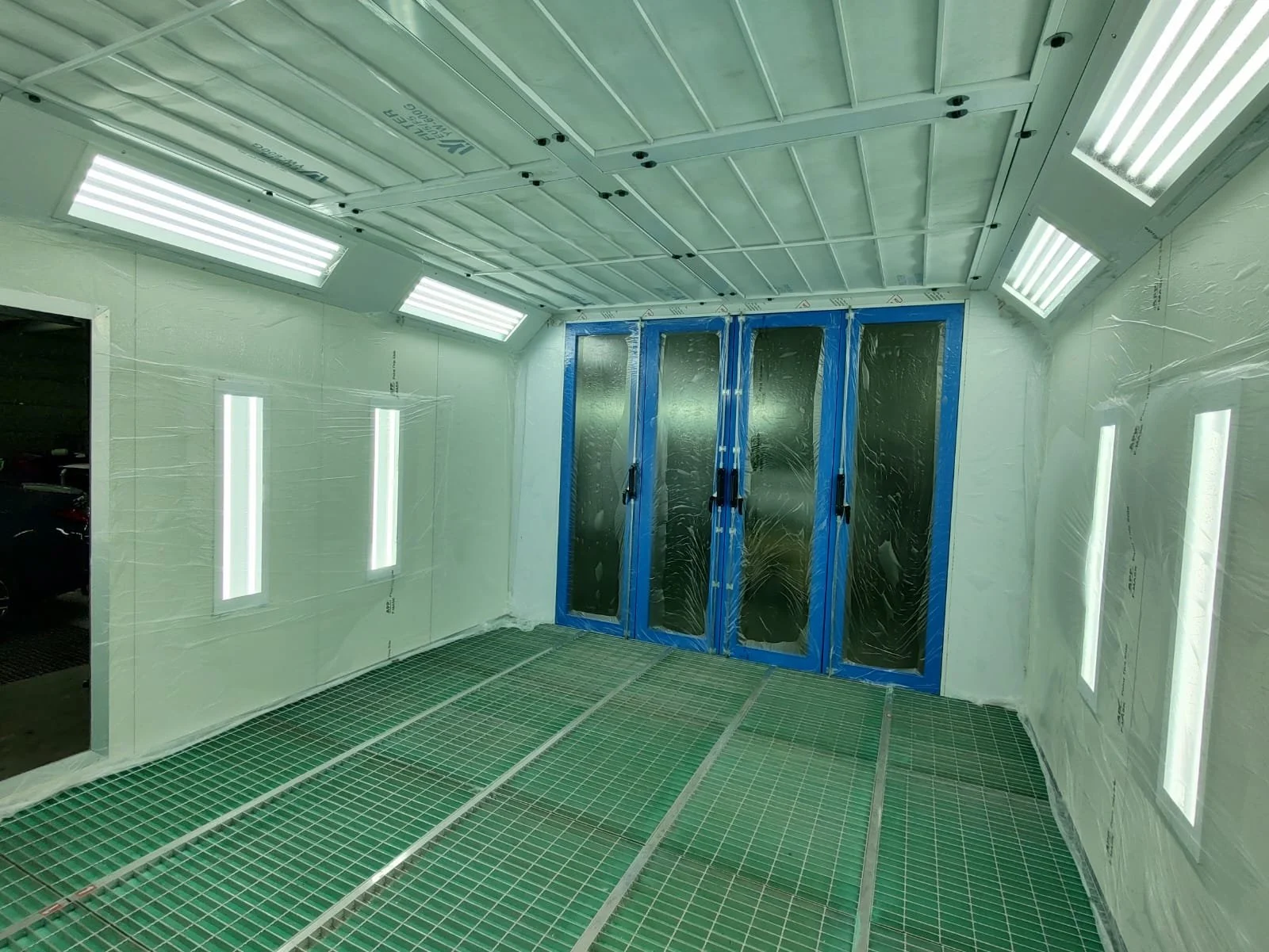 Ein Raum, der wahrscheinlich in einer Lackierwerkstatt oder einem ähnlichen Bereich liegt, mit weißen Wänden, blauen Fensterrahmen, modernen LED-Lichtleisten an der Decke und Schutzgittern auf dem Boden.