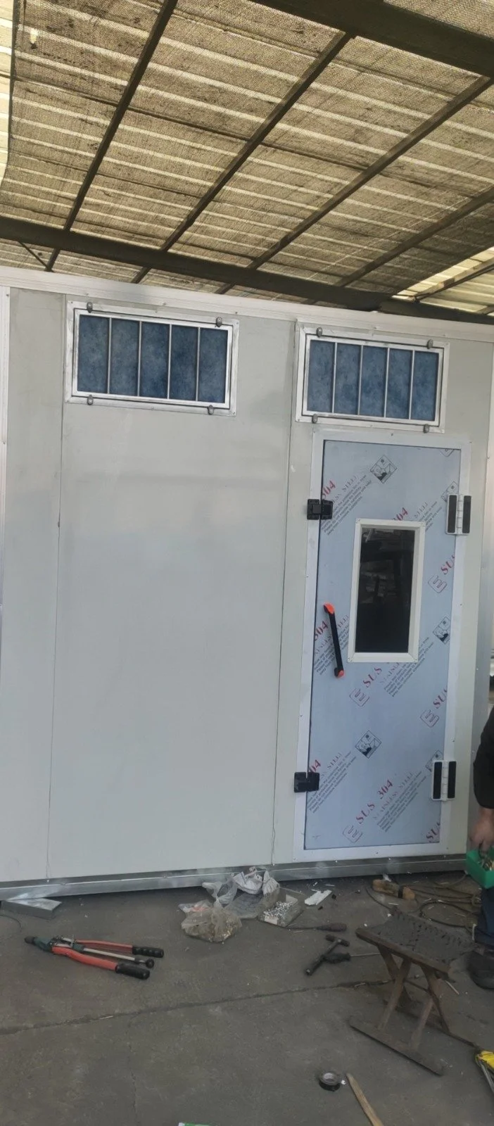 Ein Container oder eine modulare Einheit mit zwei kleinen Fenster und einer Tür, auf der Baustelle gebaut oder installiert wird.