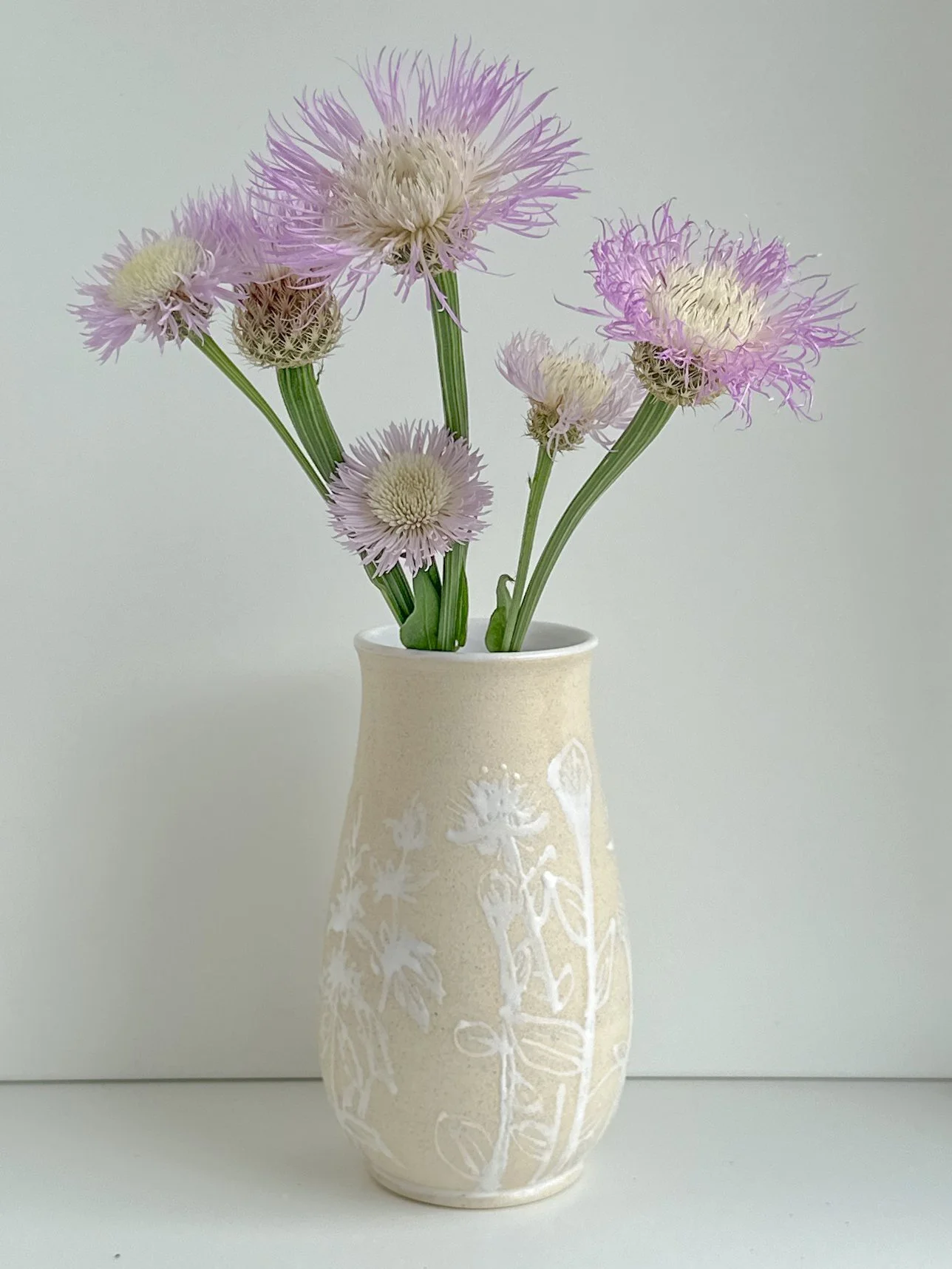 Painted beige ceramic vase with white floral design holding light purple and white flowers against a plain background.