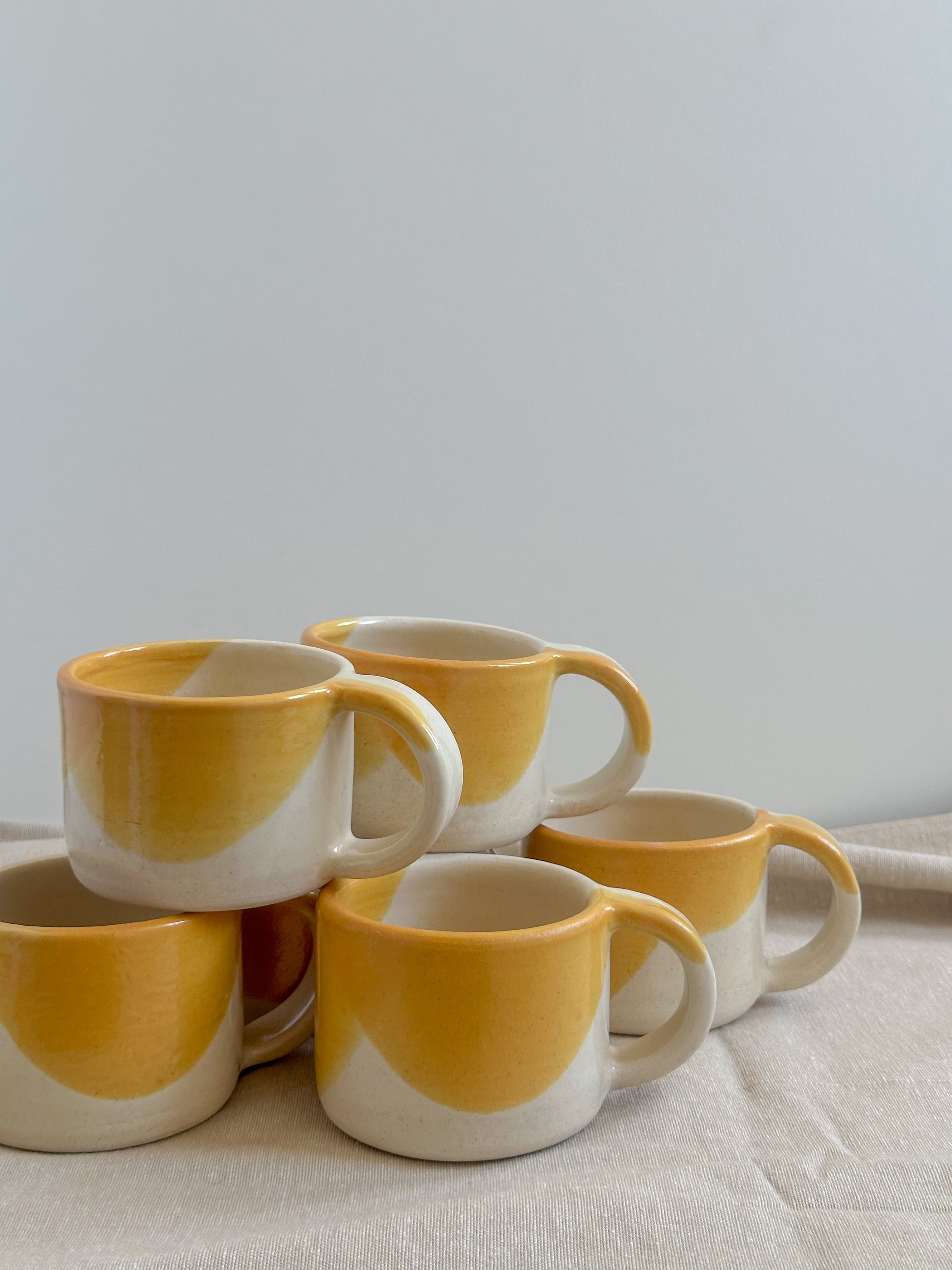 Five ceramic coffee mugs with yellow and white design, arranged in a pile on a beige cloth surface against a plain light gray background.