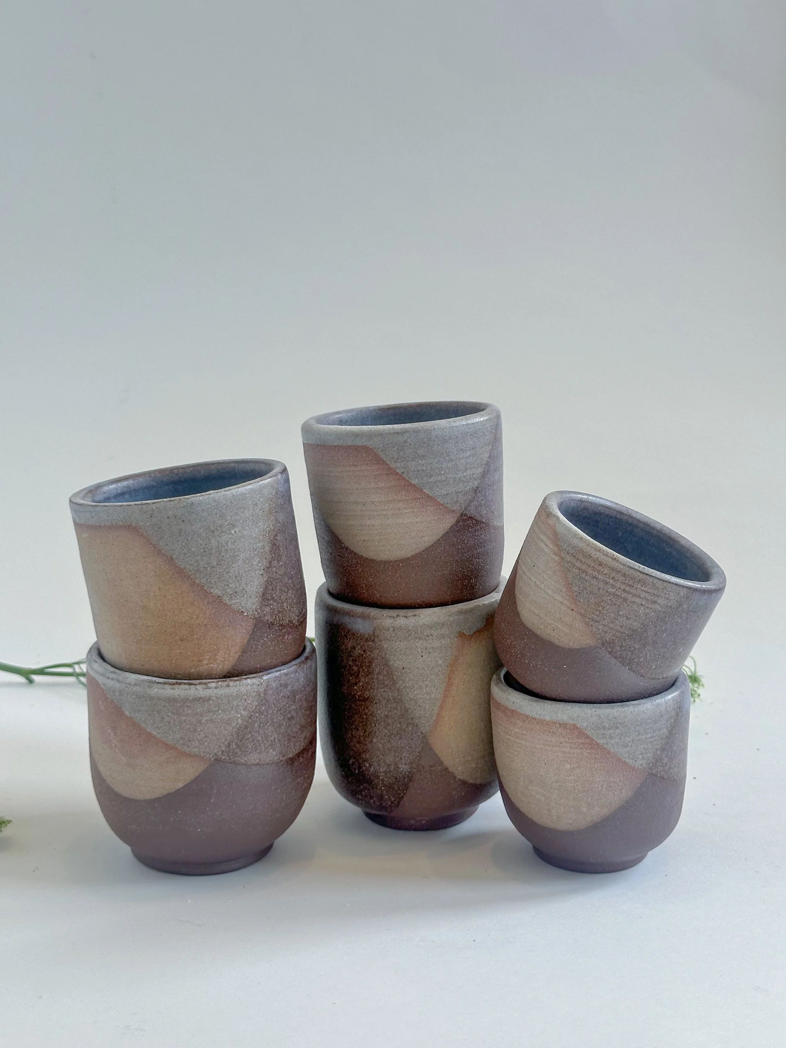 Six small ceramic cups with abstract earth-tone patterns arranged in a pyramid formation against a plain white background.