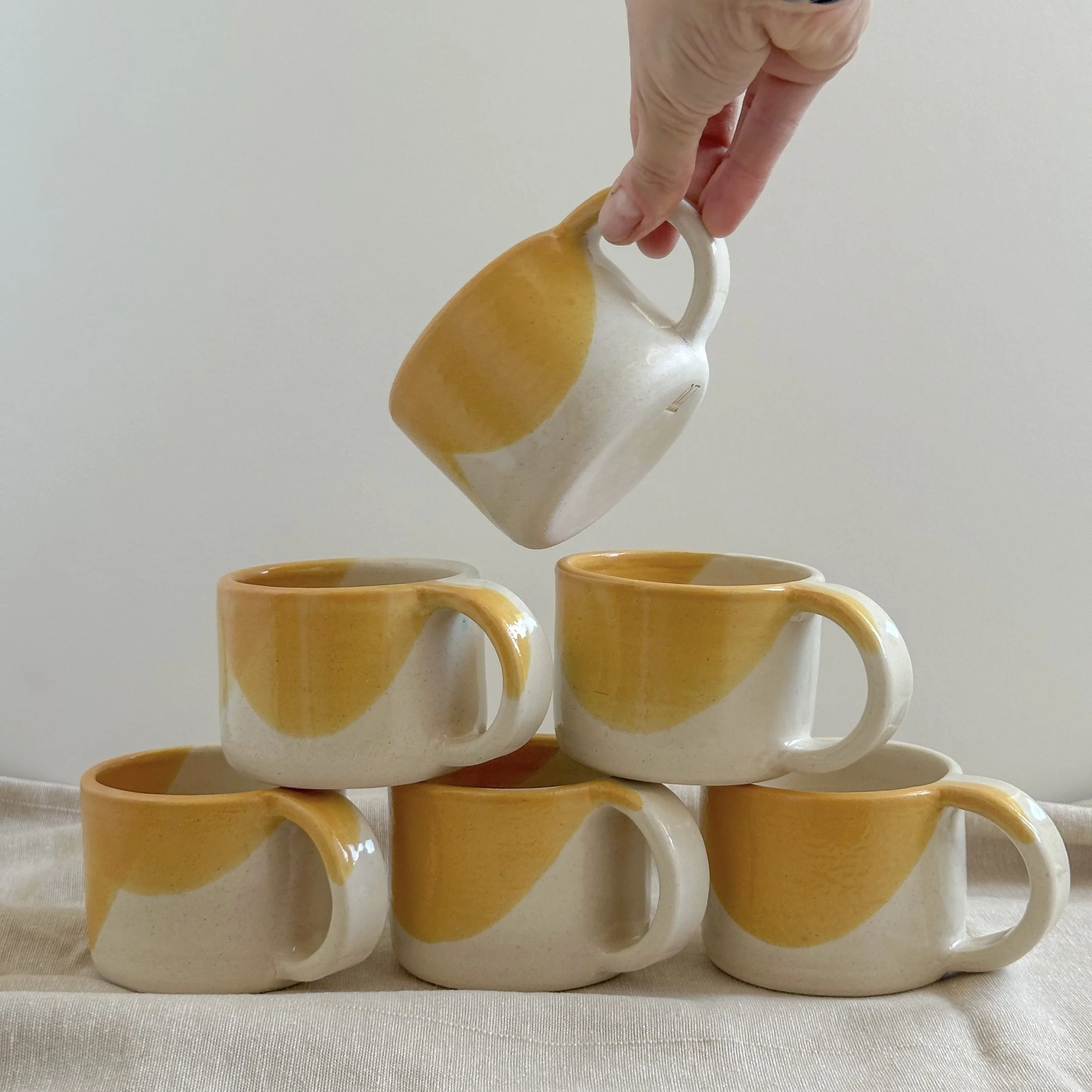 A person stacking six yellow and white ceramic coffee mugs with a yellow design against a plain background.