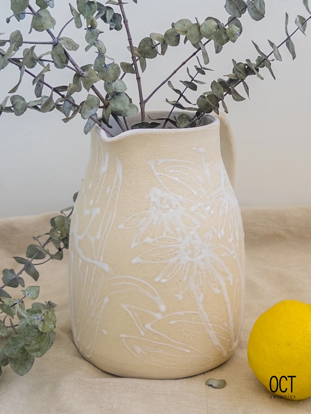 Decorative beige ceramic vase with white floral and leaf patterns, filled with eucalyptus branches, placed on a beige fabric surface next to a yellow lemon.