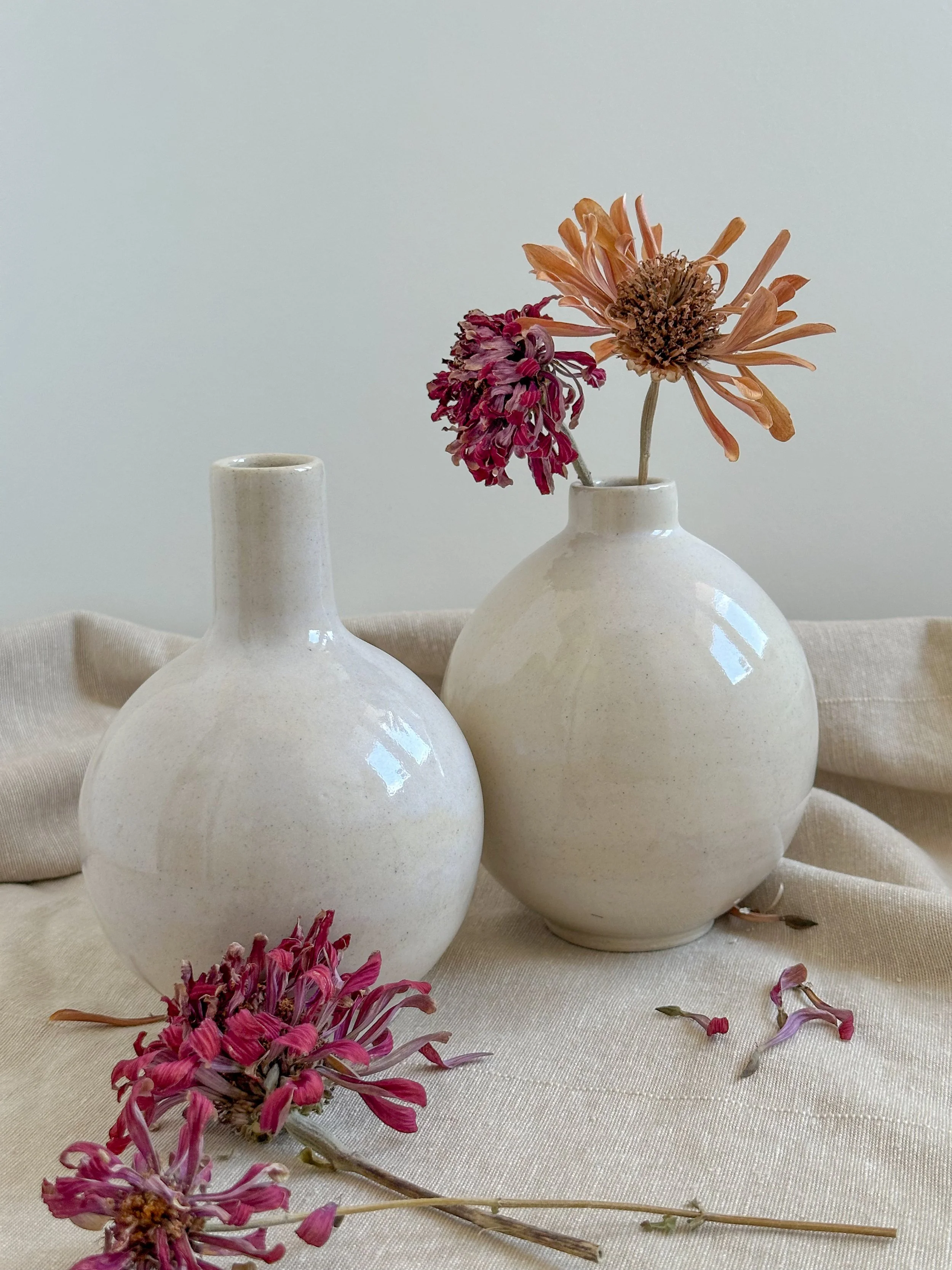 Two cream-colored ceramic vases, one with dried pink and orange flowers, placed on a light-colored fabric surface with some fallen petals.