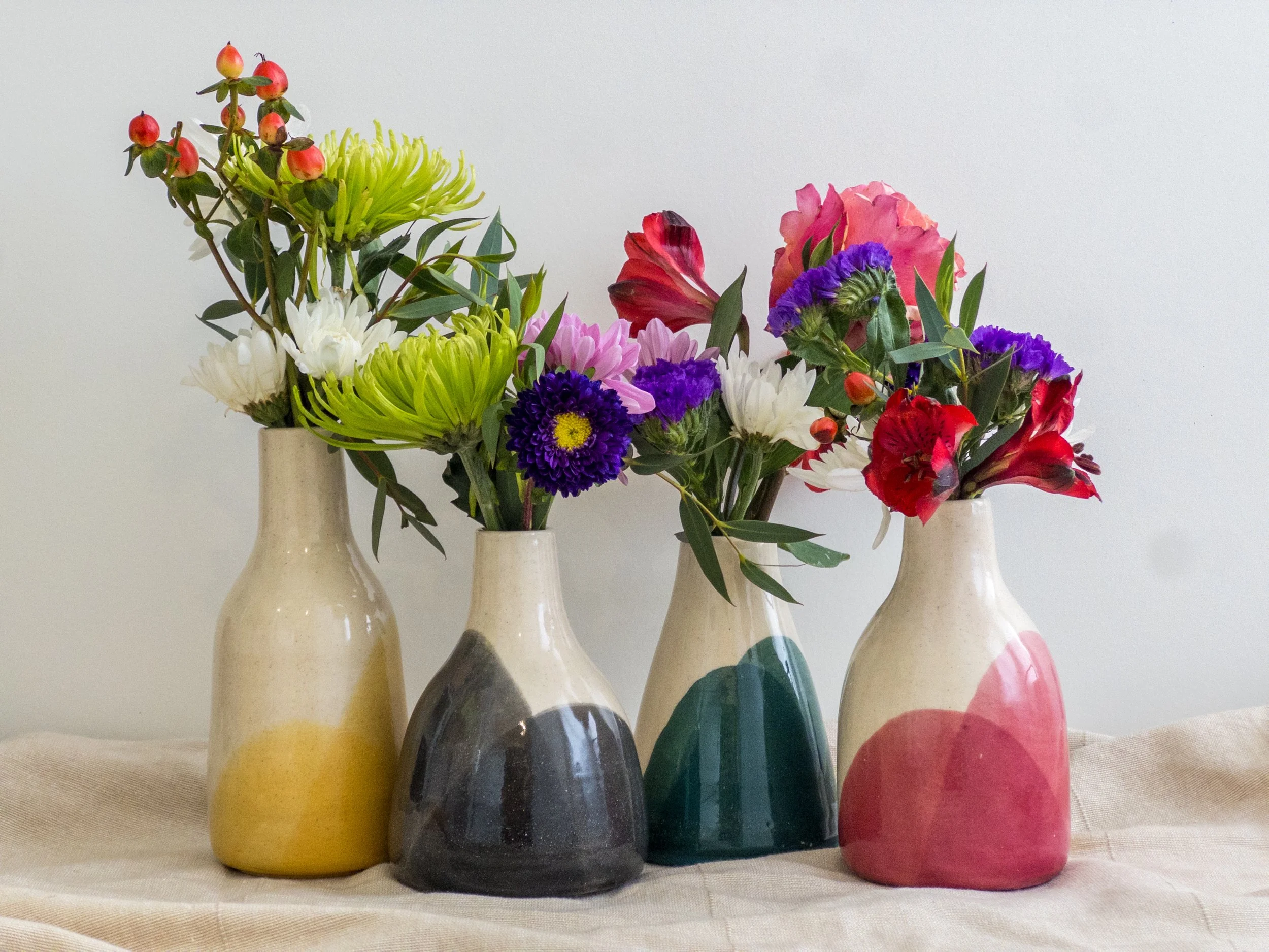 Four ceramic vases with colorful flowers on a beige cloth, against a plain white wall.
