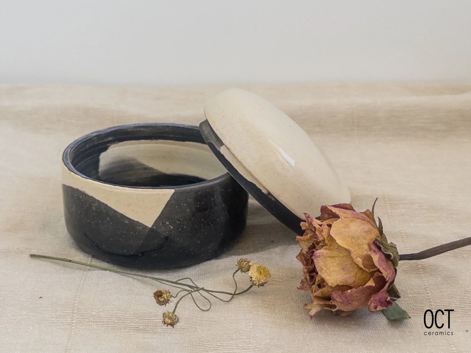 A black and beige glazed ceramic container with a lid leaning to the side, positioned on a light fabric surface alongside a dried orange and yellow flower and small dried buds.