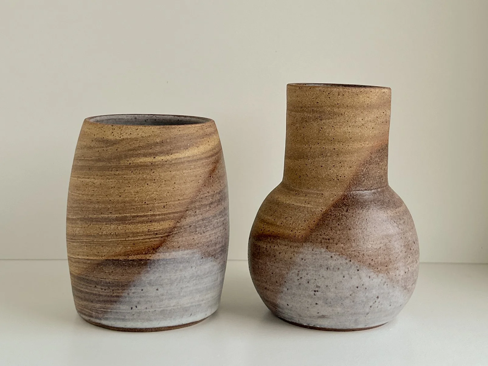 Two handcrafted ceramic vases with earthy tones and wavy pattern, one shorter and wider, the other taller and narrower, placed on a white surface against a plain background.