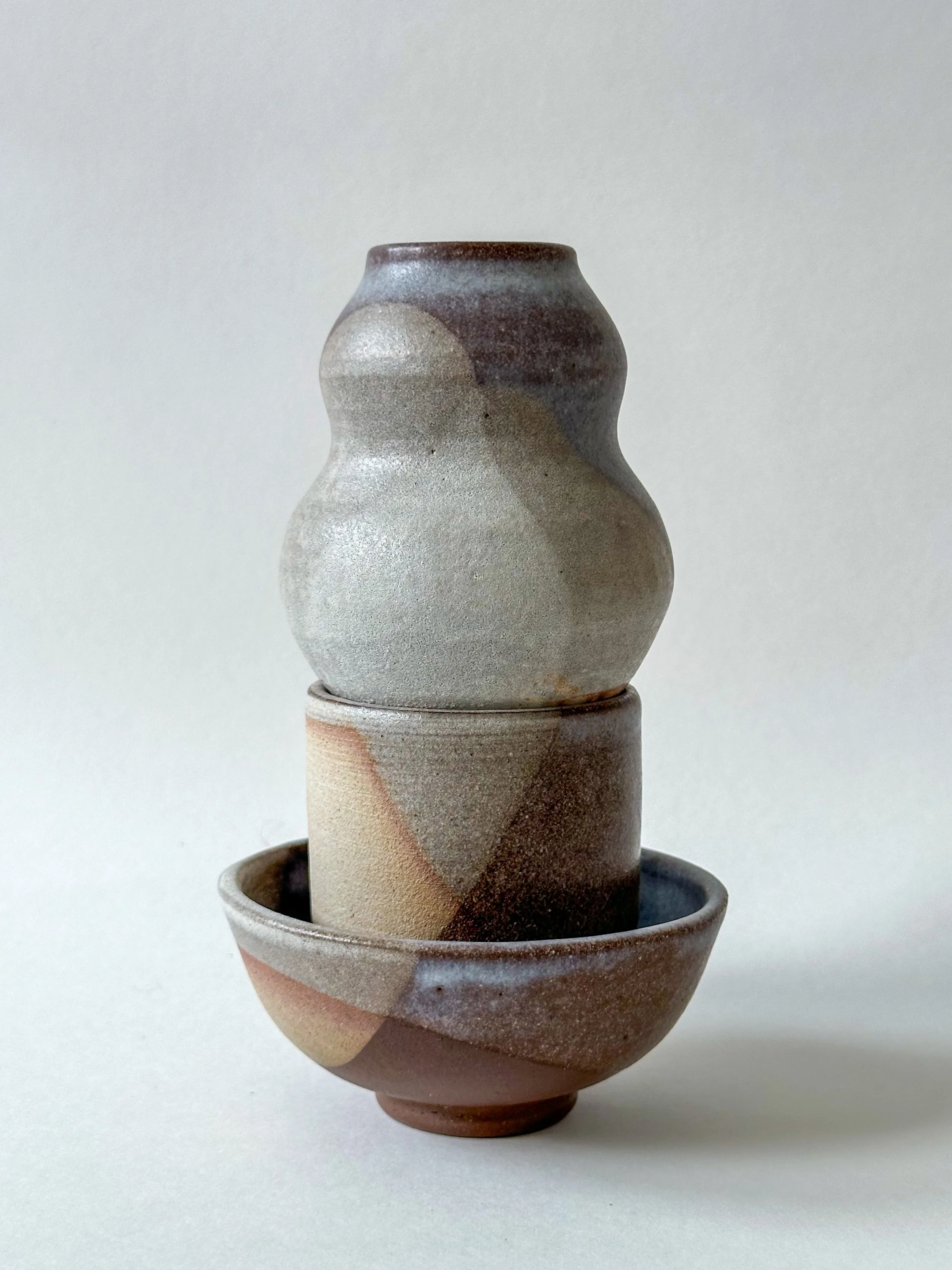 Two stacked ceramic vases and a bowl with abstract earthy-colored glaze, minimalistic and textured, against a plain white background.