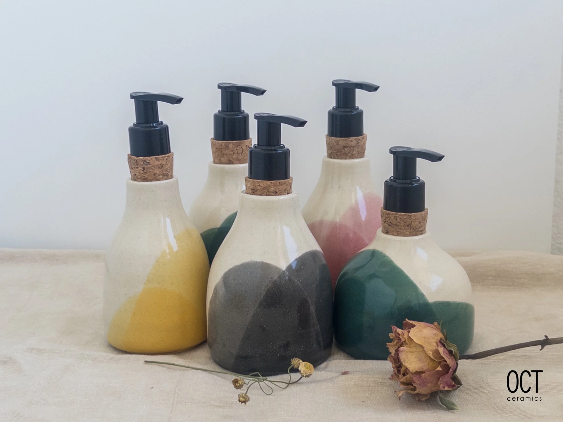 Six handmade ceramic soap dispensers with pumps, featuring colorful abstract designs, arranged on a light surface with dried flowers nearby.