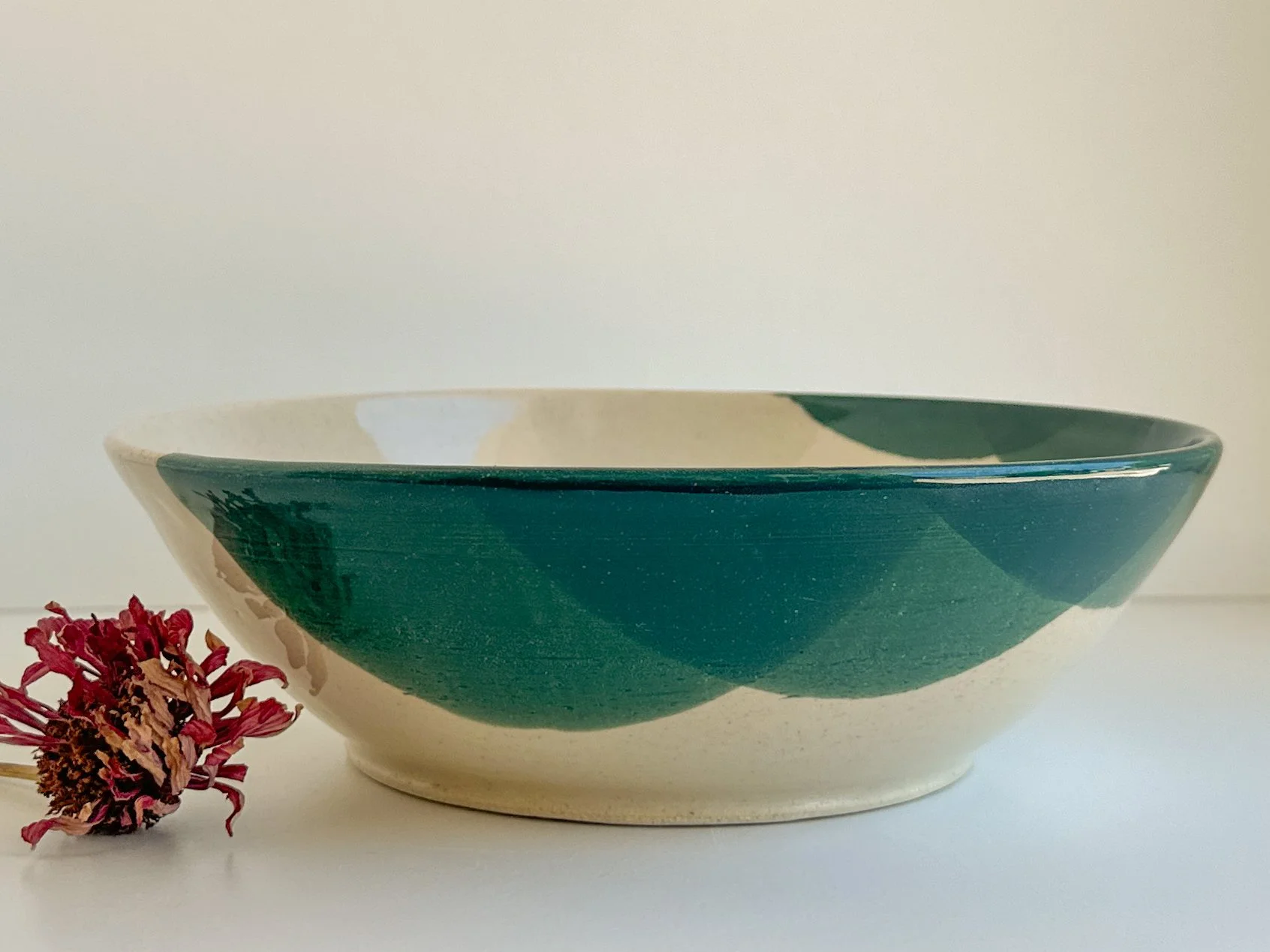 A ceramic bowl with a green and beige pattern, resting on a white surface next to a withered red flower.