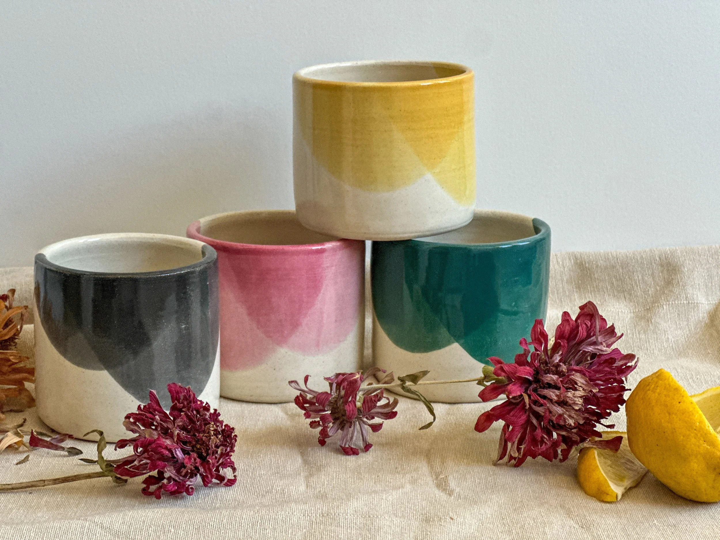 Five colorful ceramic mugs stacked with three on the bottom and two on top, surrounded by pink dried flowers and a sliced lemon, on a neutral cloth surface.
