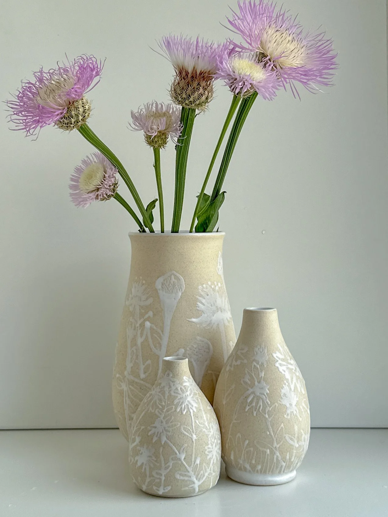 A set of three white vases with floral patterns, with purple and white flowers in the tallest vase against a plain background.