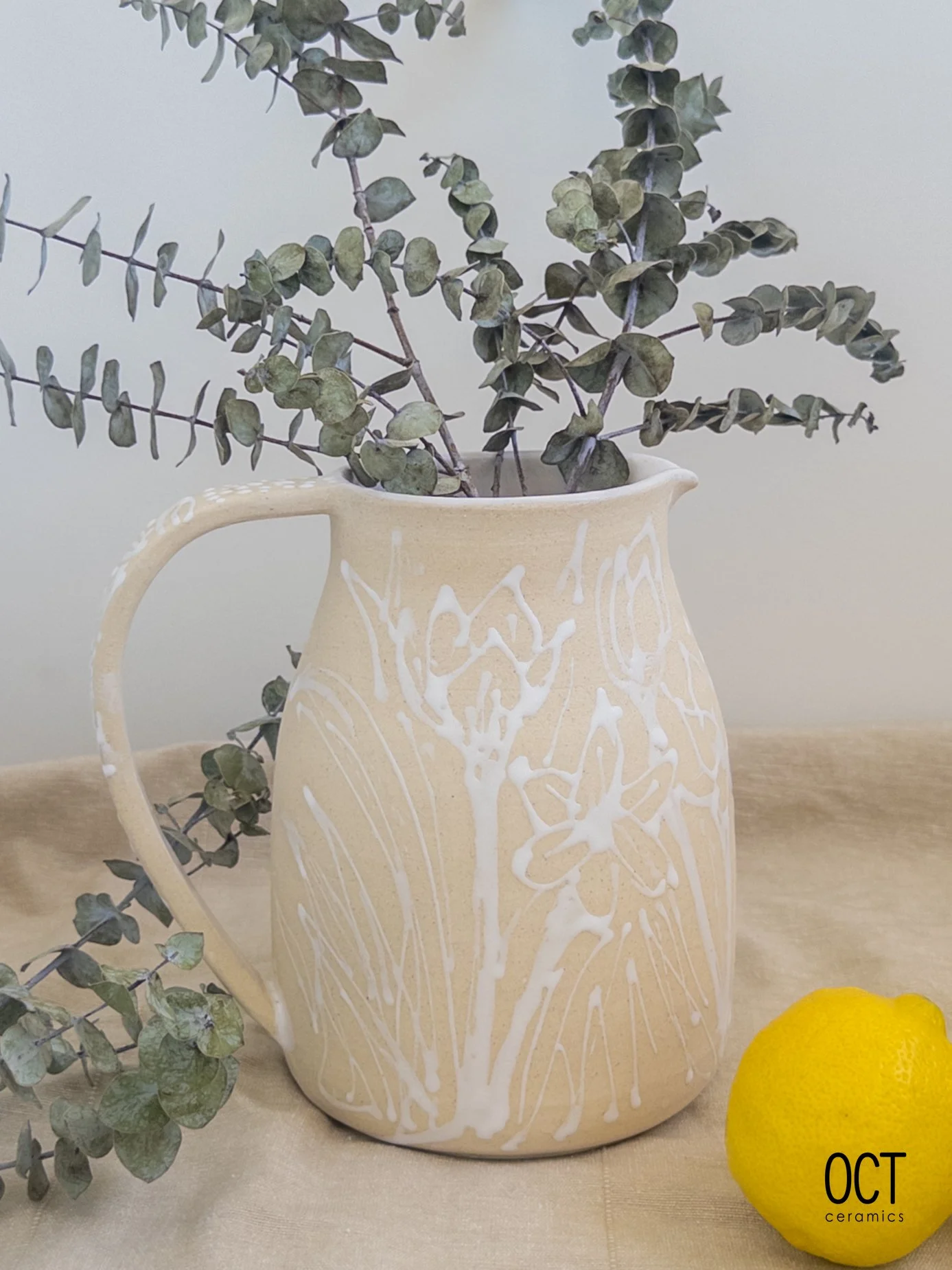 A beige ceramic pitcher with white floral patterns, holding eucalyptus branches, placed on a beige cloth with a yellow lemon next to it. The logo 'OCT ceramics' is in the bottom right corner.