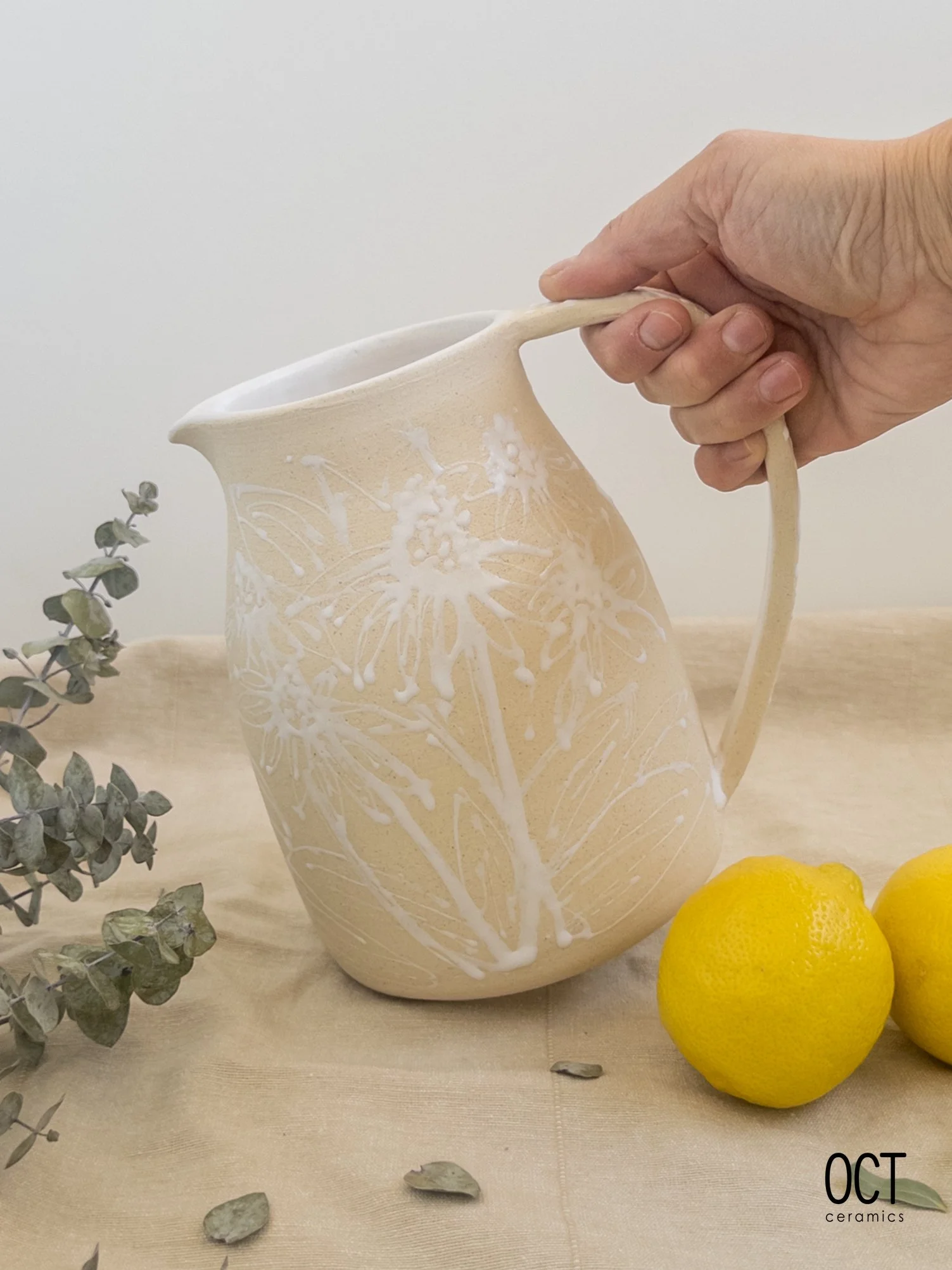Hand holding a beige ceramic pitcher with white floral and leaf design, next to two lemons and eucalyptus leaves on a beige table.