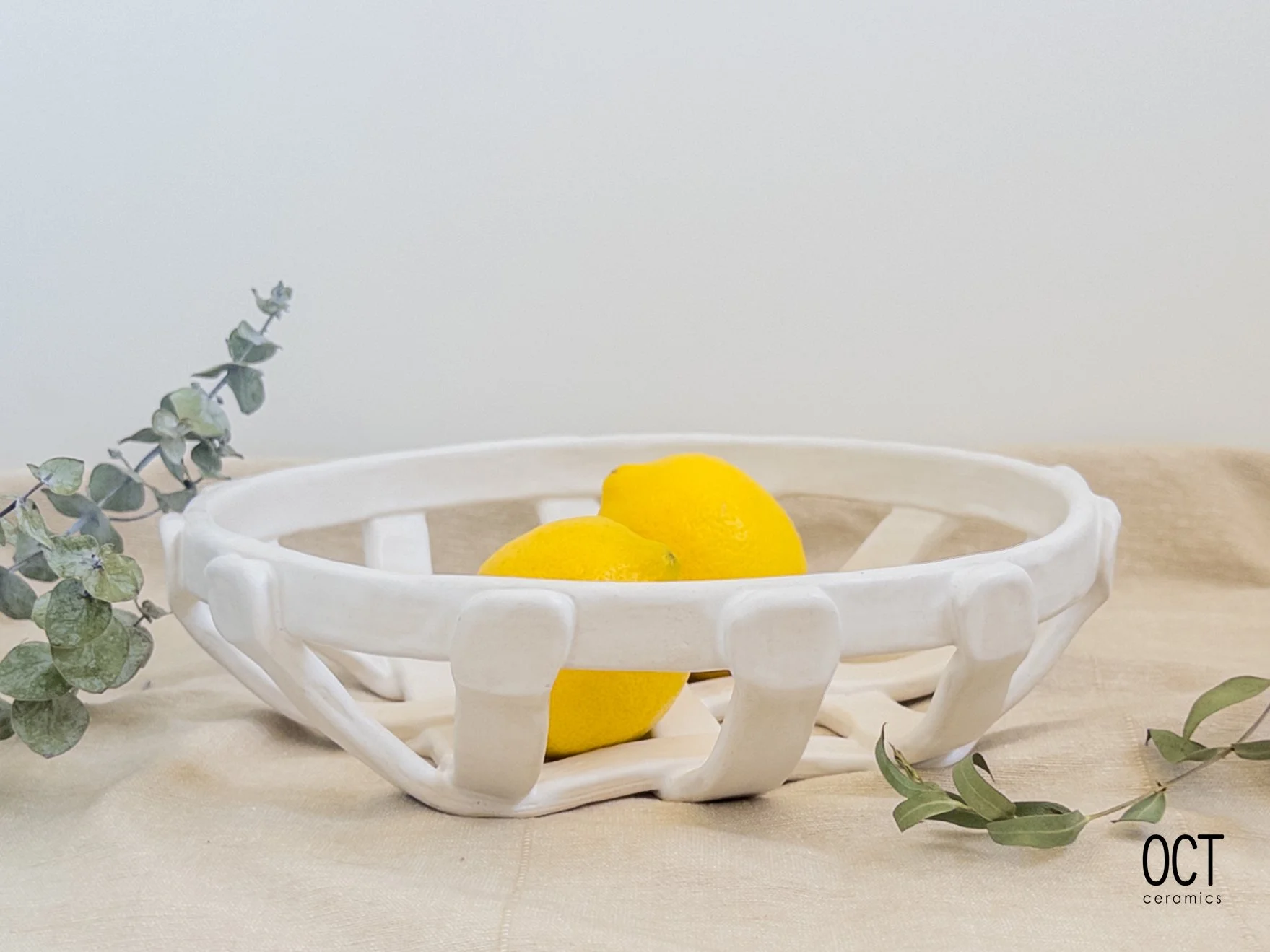 White ceramic bowl with open lattice design holding two lemons, placed on a beige fabric surface with green leafy branches on either side, against a plain light background.
