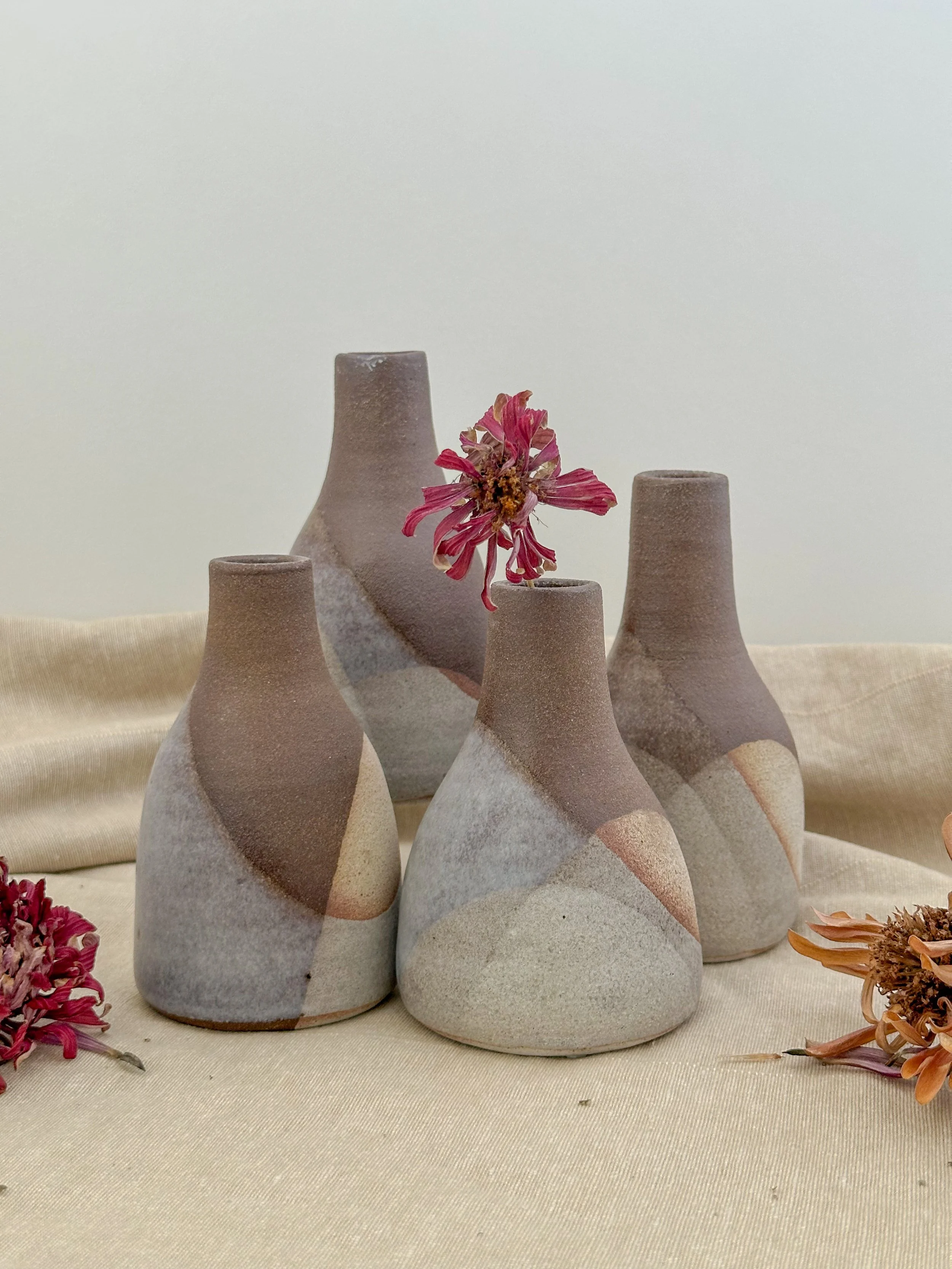 Four ceramic vases with abstract design and a pinkish flower in one vase, arranged on a beige fabric surface with dried flowers around.