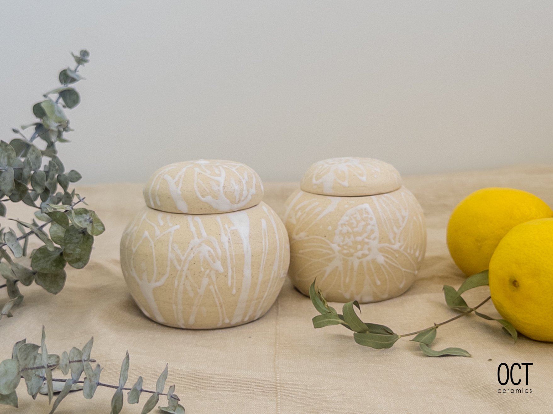Two beige ceramic jars with white abstract patterns, next to yellow lemons and eucalyptus leaves on a light-colored surface.