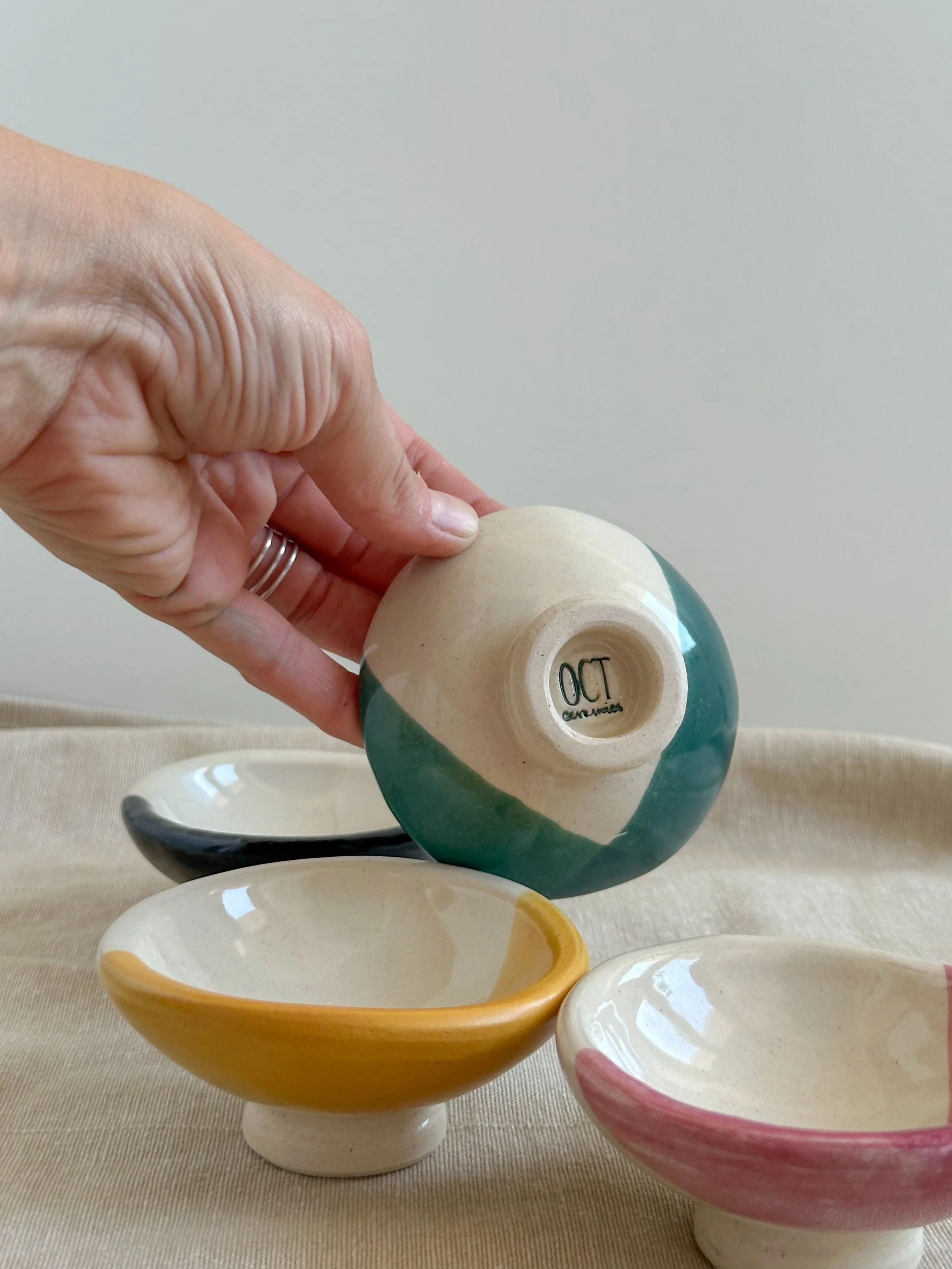 A hand holding a small, colorful ceramic bowl upside down with three other small ceramic bowls on a beige fabric surface.