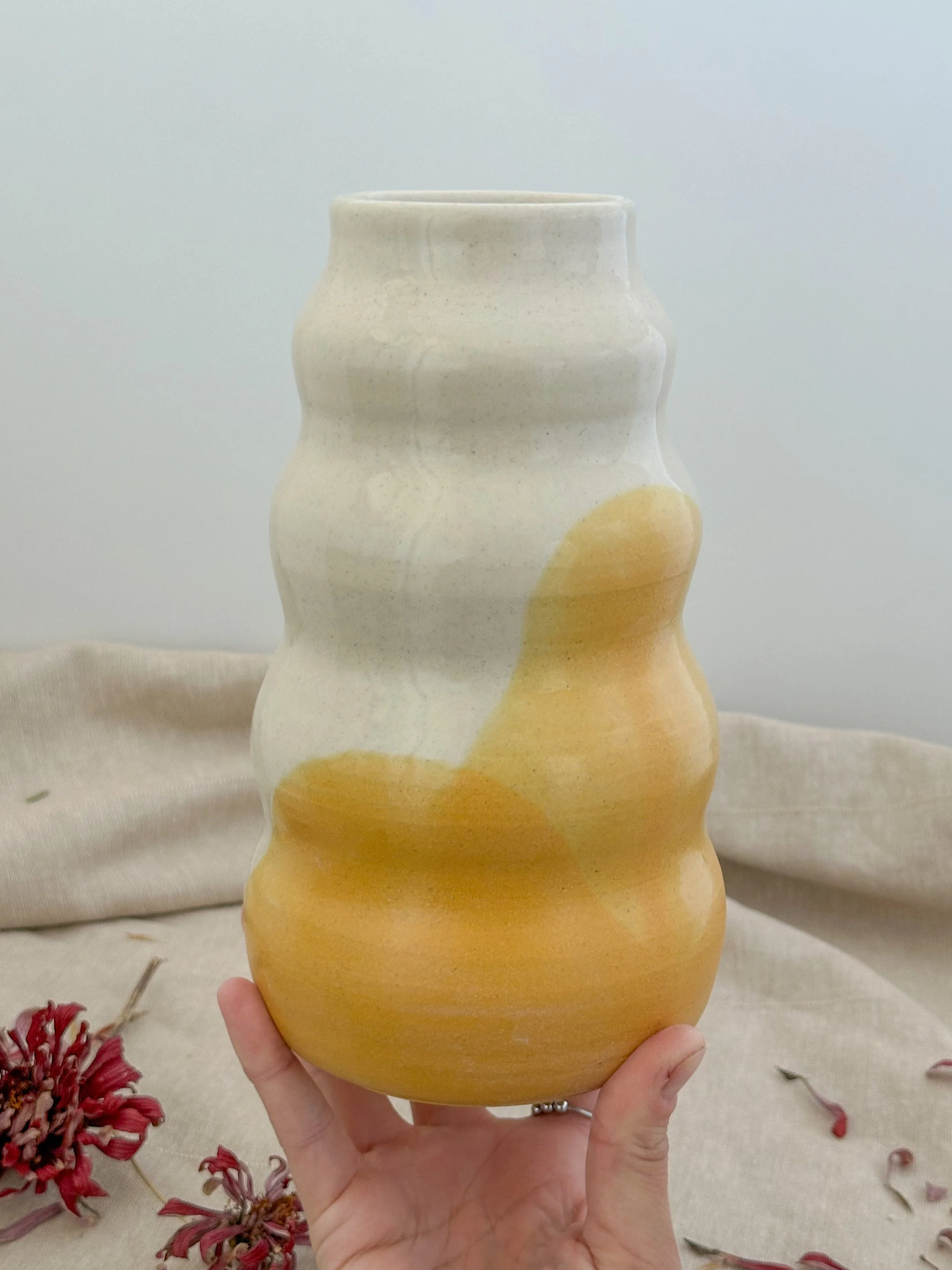 A person holding a ceramic vase with a wavy, textured surface, featuring a gradient of soft white to warm yellow tones, against a neutral background with dried flowers on a fabric surface.