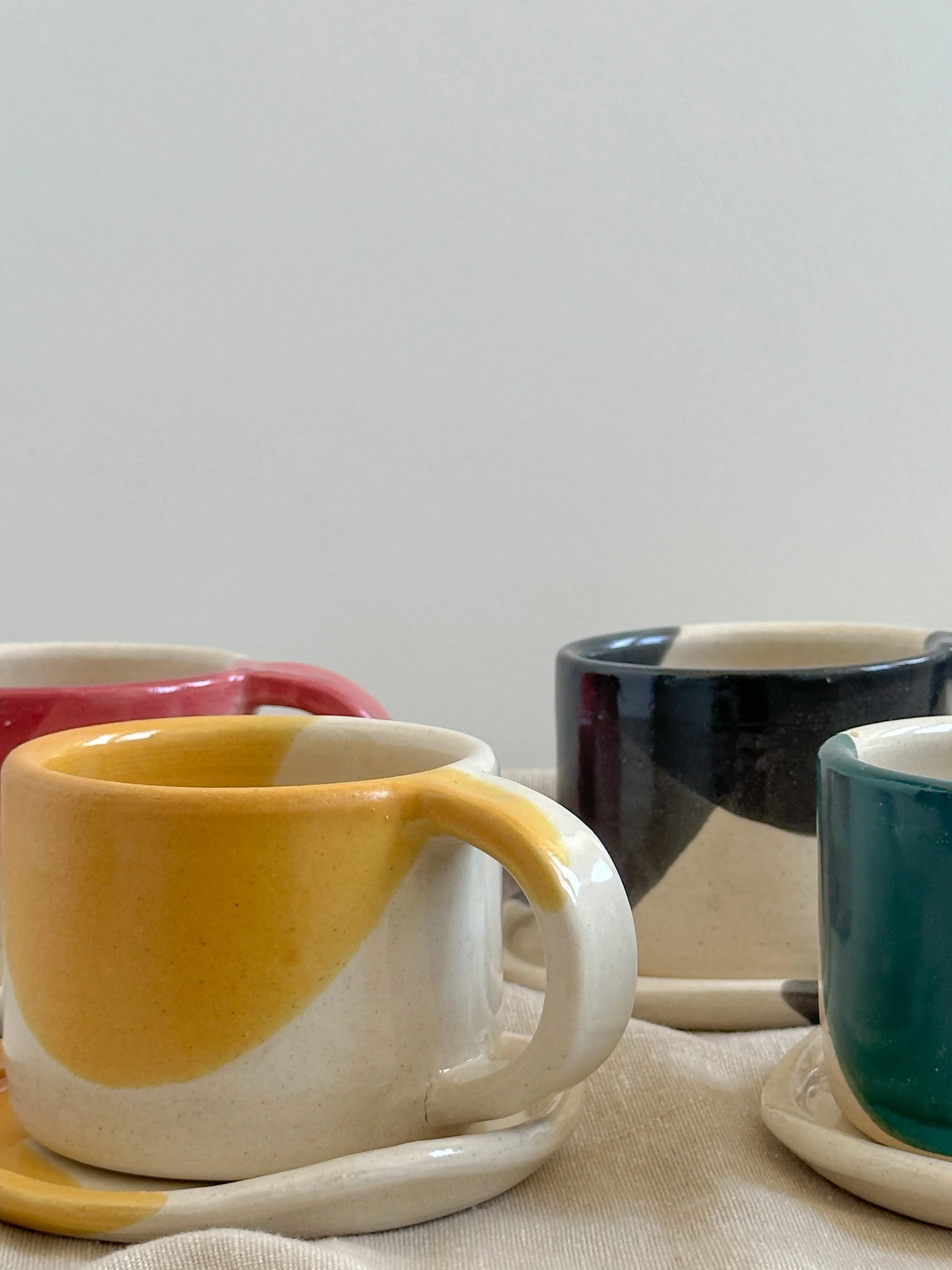 Four colorful ceramic coffee mugs on matching saucers, arranged on a beige cloth, with a plain white background.