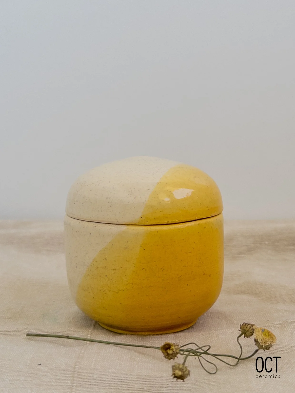 A yellow ceramic container with a rounded shape, placed on a cream-colored cloth with dried flowers in front, and a plain light background.
