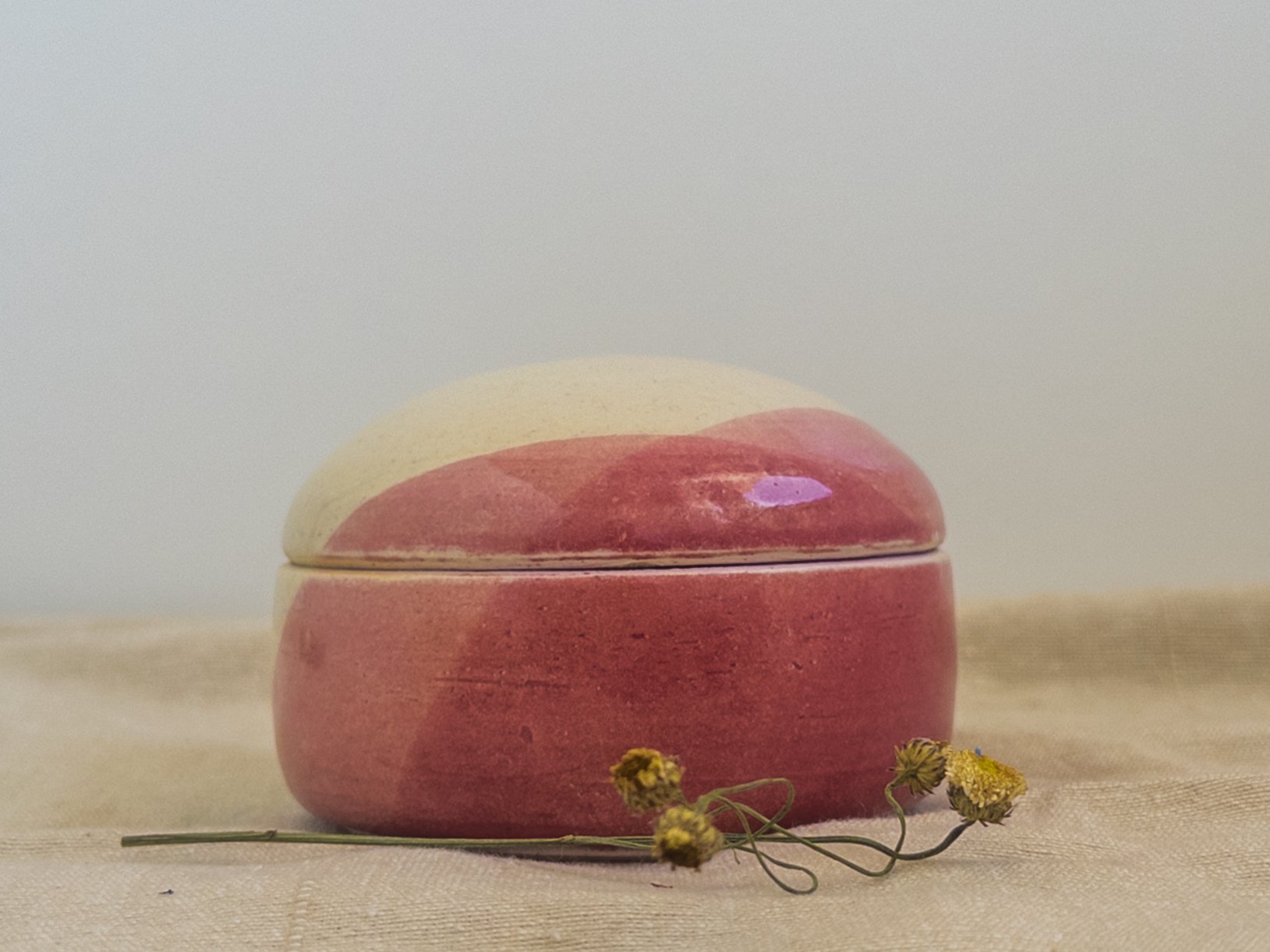 A pink and white ceramic container with a lid, placed on a beige cloth surface, accompanied by small dried yellow flowers.