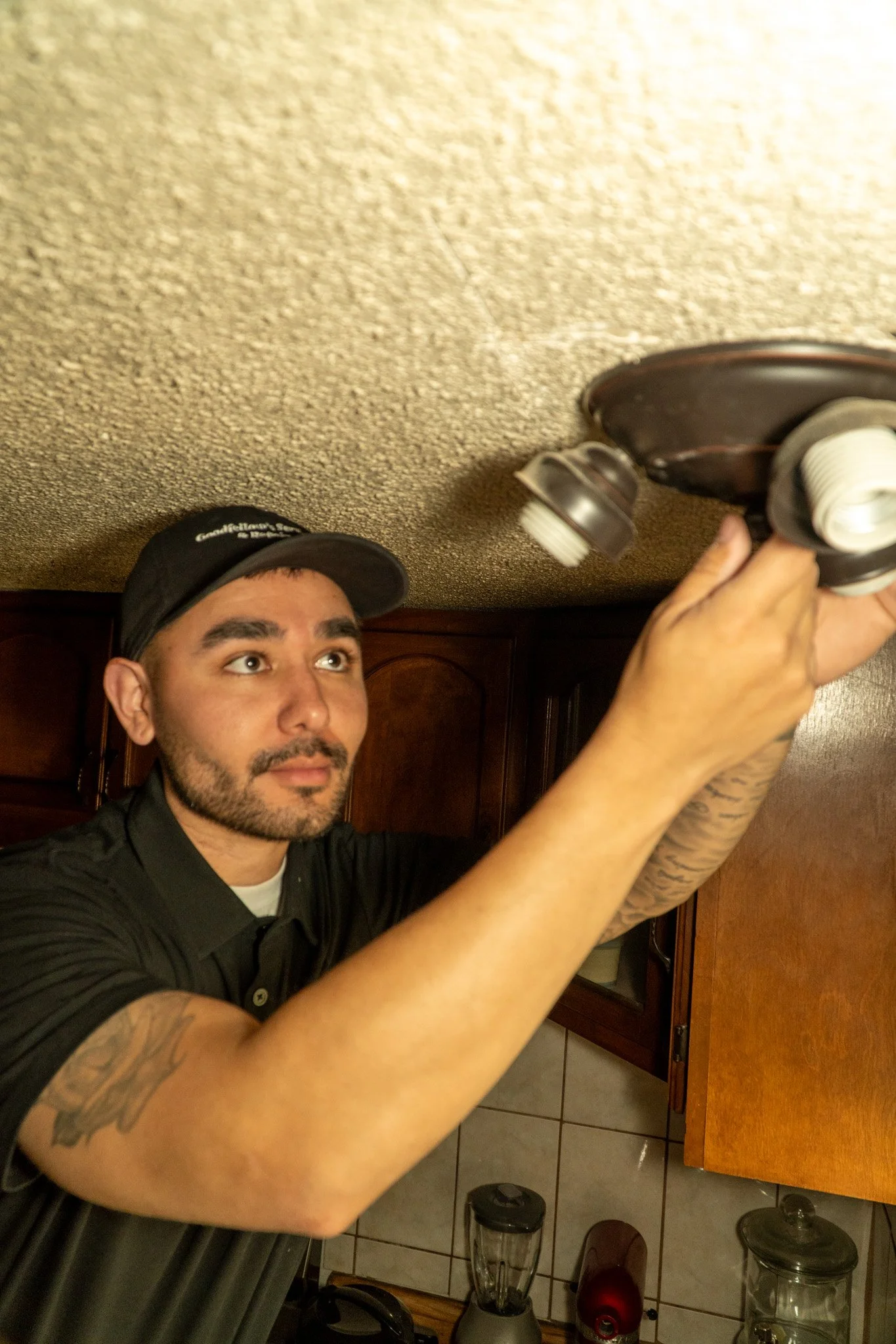 A man installing a ceiling light fixture in a kitchen, using a screwdriver, with wooden cabinets and Kitchen appliances in the background.