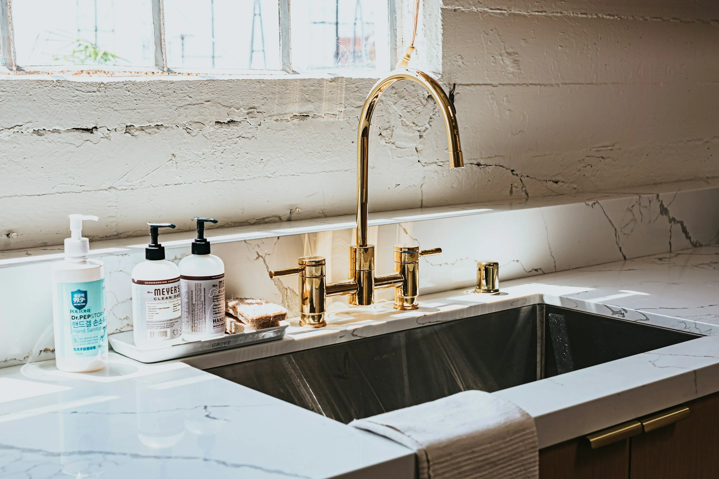 A modern kitchen sink area with white marble countertops, a gold faucet, two black soap dispensers, a bottle of hand sanitizer, and a sponge on a tray. Sunlight streams through a window above, illuminating the scene.