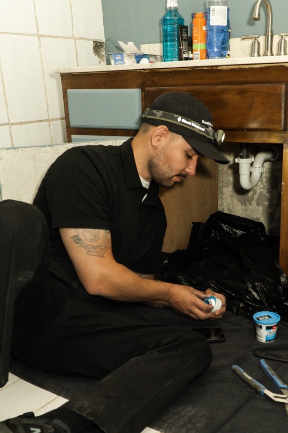Man sitting on the floor under a kitchen sink, working on plumbing with tools, wearing a headlamp and a black shirt.