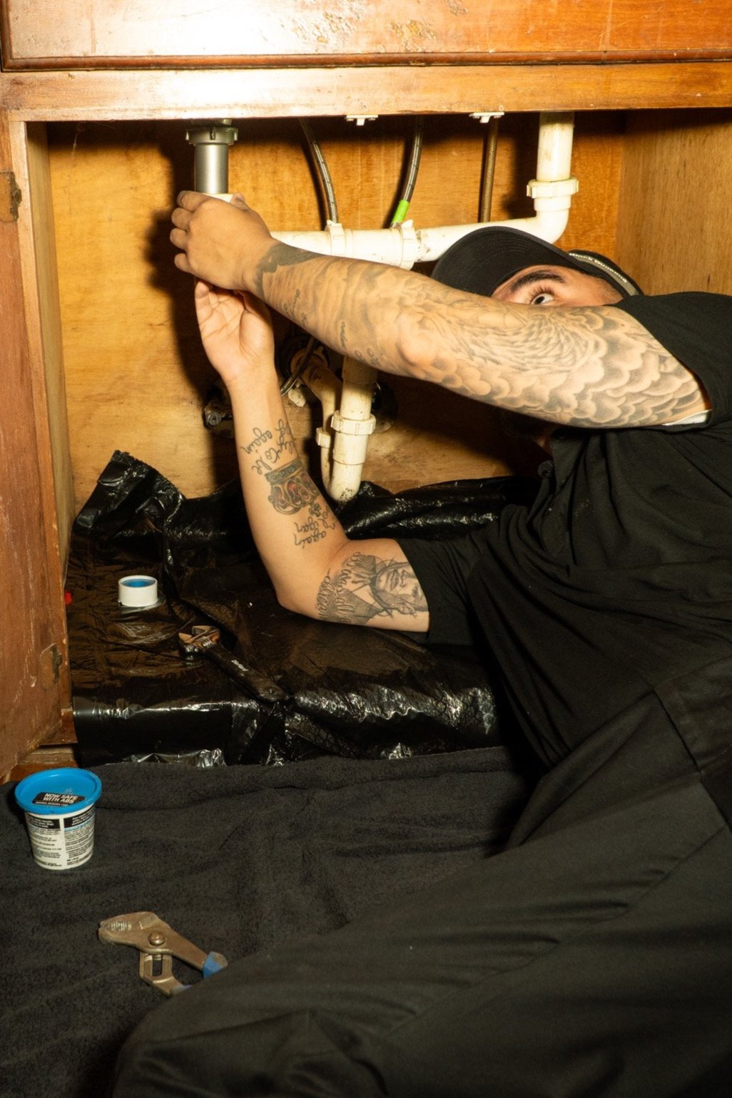 A man with tattoos working under a kitchen sink, fixing pipes.