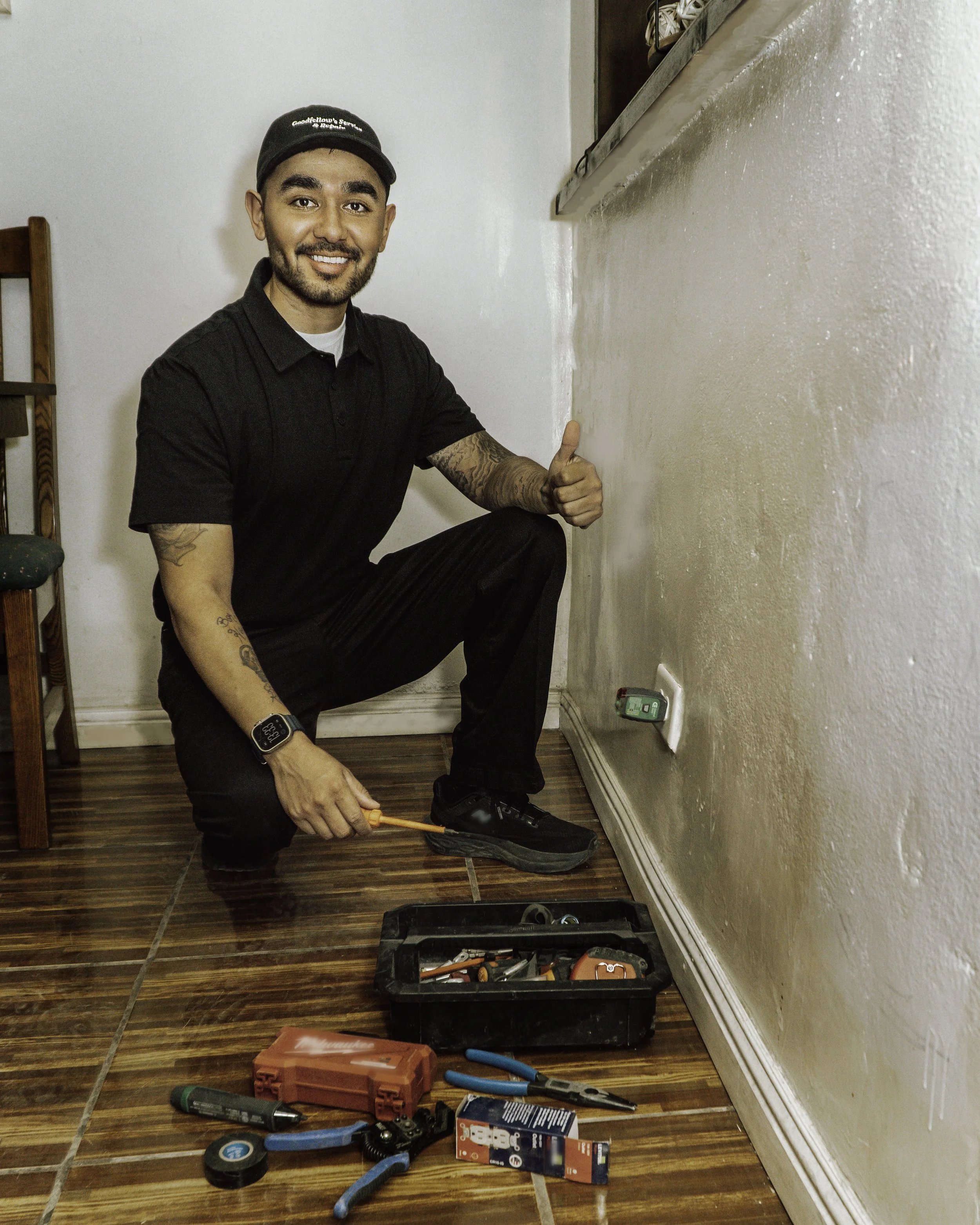 A man crouching on a hardwood floor, giving a thumbs up, during a home electrical repair. He is holding a screwdriver, with tools and a toolbox nearby, and an open electrical outlet on the wall.