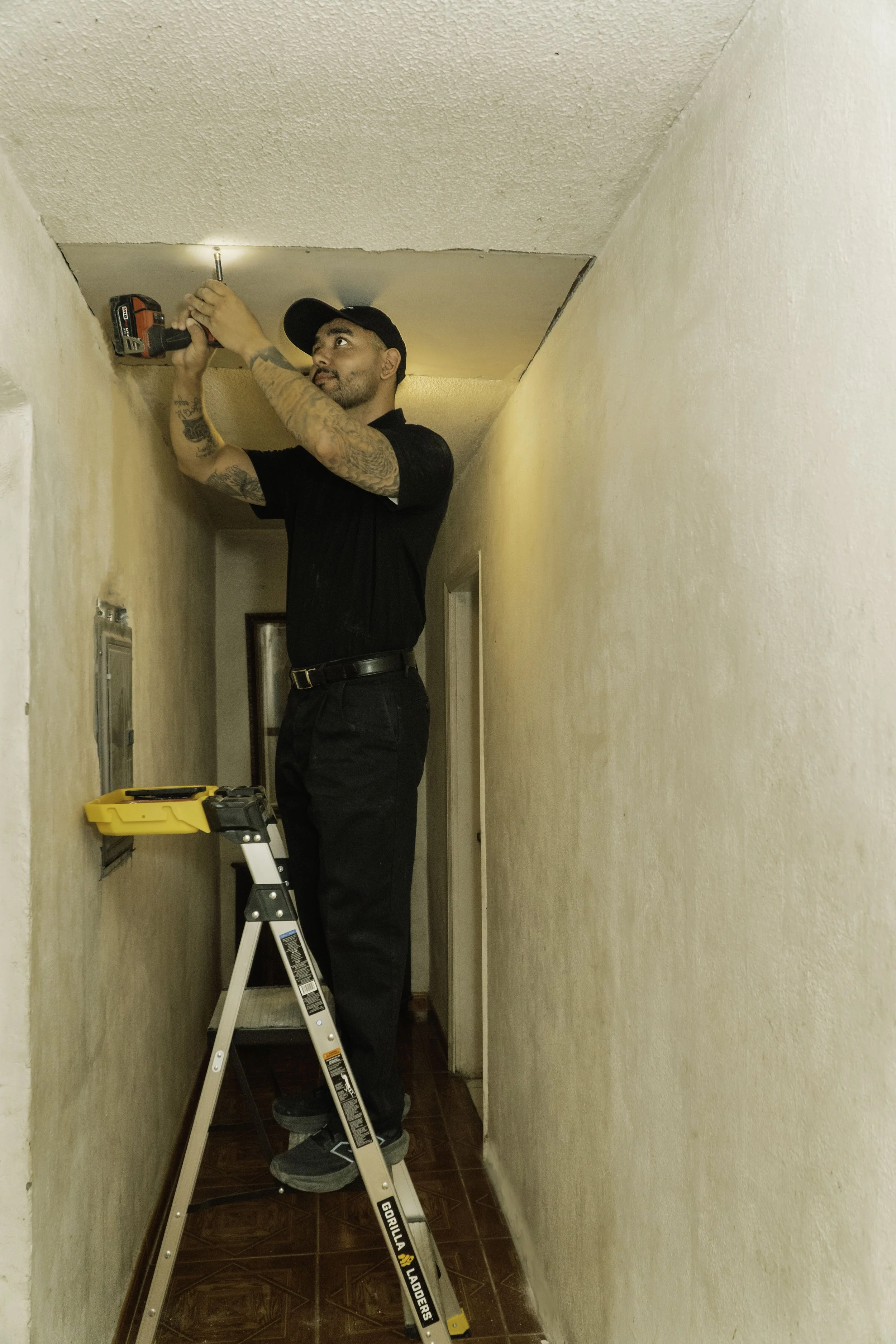 Man on a ladder installing a ceiling light fixture in a narrow hallway.