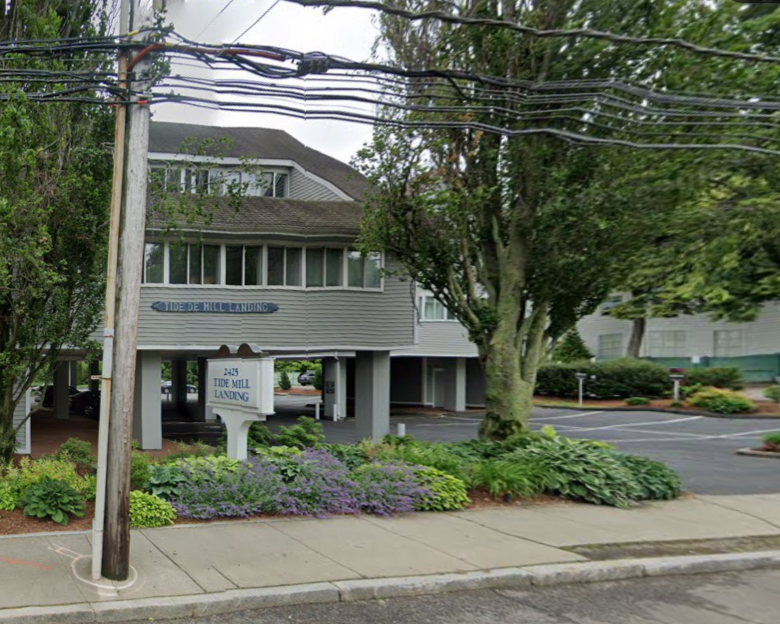 A building with a parking garage underneath, labeled 'Tide Mill Landing,' surrounded by landscaped bushes and trees. A street lamp pole and power lines are visible in the foreground.