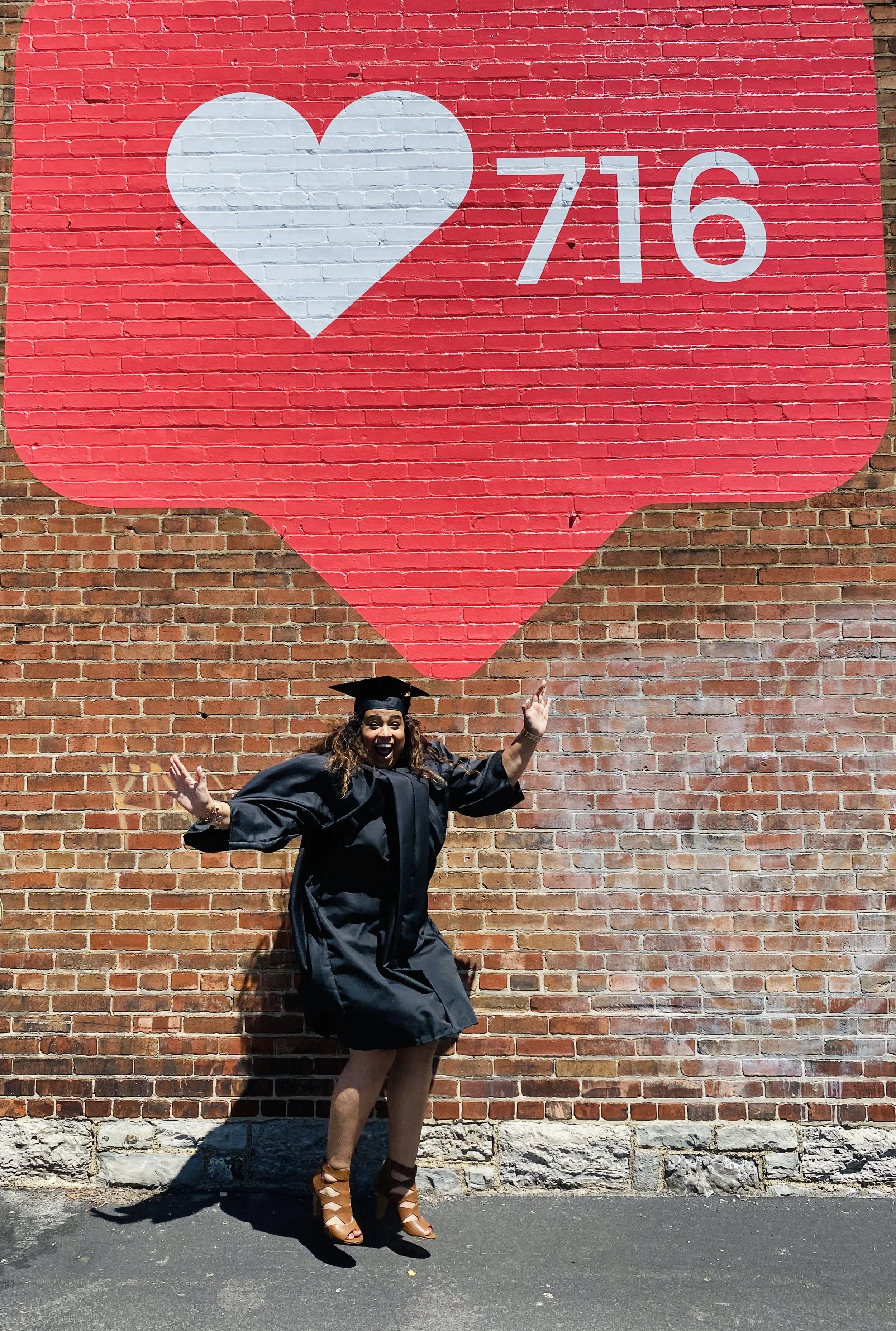 Person in graduation gown jumping in front of brick wall with large red heart and number 716 painted on it.