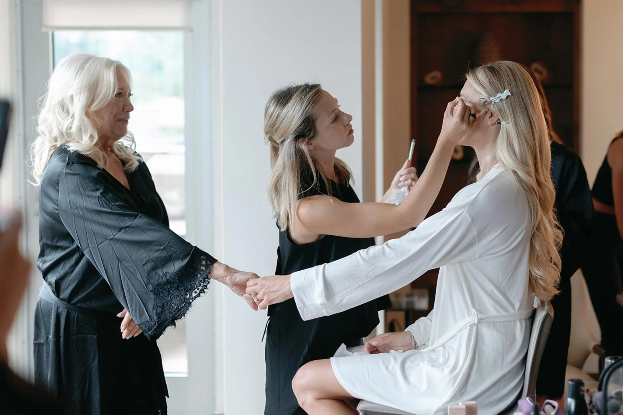 Bride getting makeup done, holding hands with her mom, in brightly lit room.