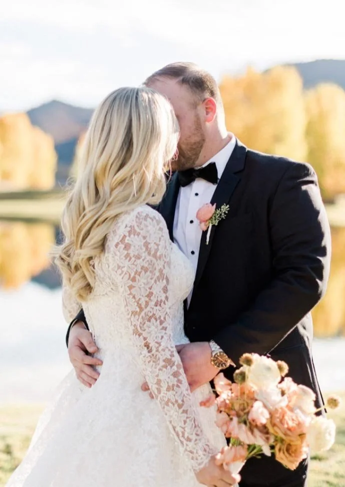 A bride and groom kissing outdoors during a wedding, with the bride in a lace dress holding a bouquet, and the groom in a tuxedo.