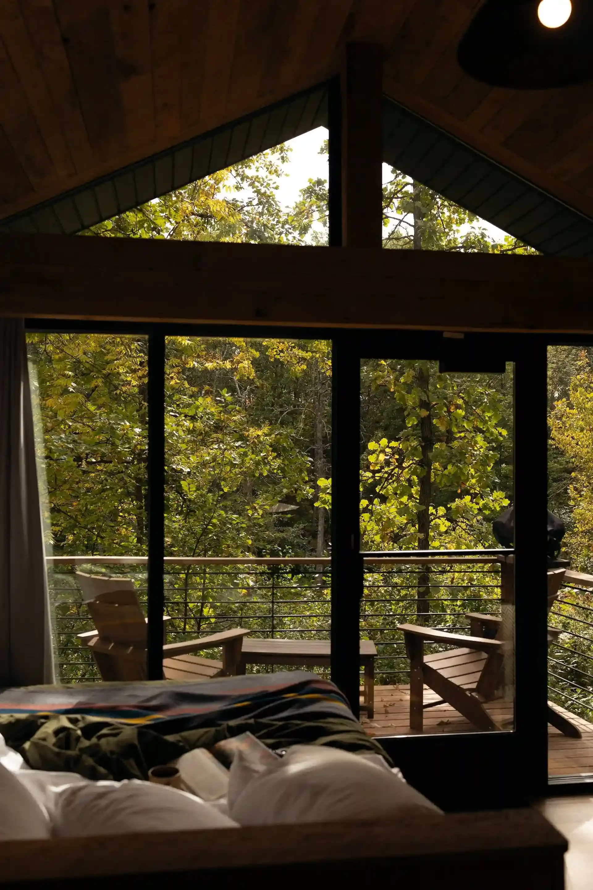 A cozy bedroom interior with a large window view of a forest. The room has a wooden ceiling and a bed with pillows and a blanket. Outside the window, there's a deck with wooden chairs and a table under trees with green and yellow leaves.