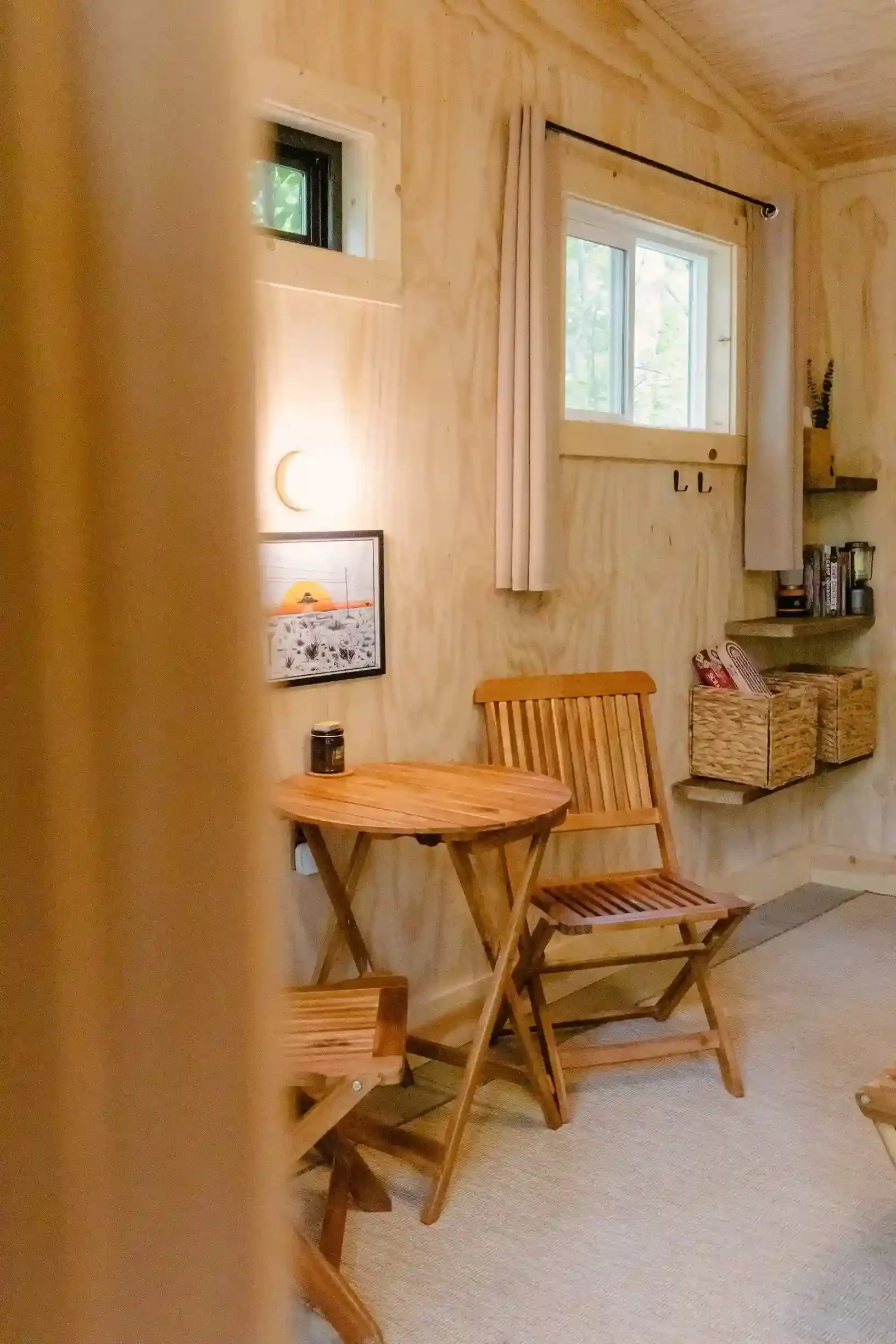 Cozy wooden room interior with a small round table and two folding chairs, a window with curtains, a framed picture on the wall, and baskets on shelves containing books and items.