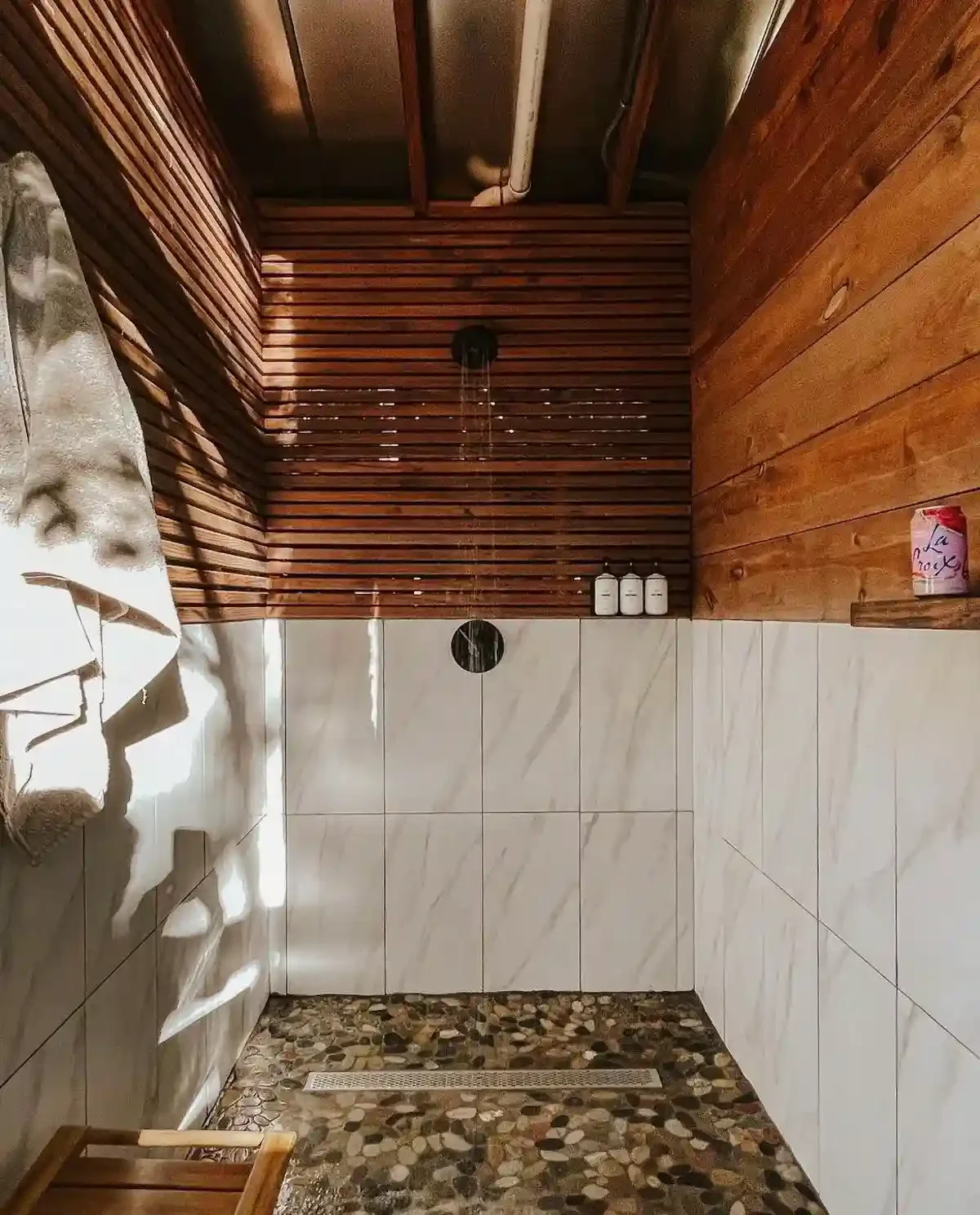 Modern rustic shower with wood paneling, stone floor, and marble walls, featuring a black shower head and small shelves with toiletries.