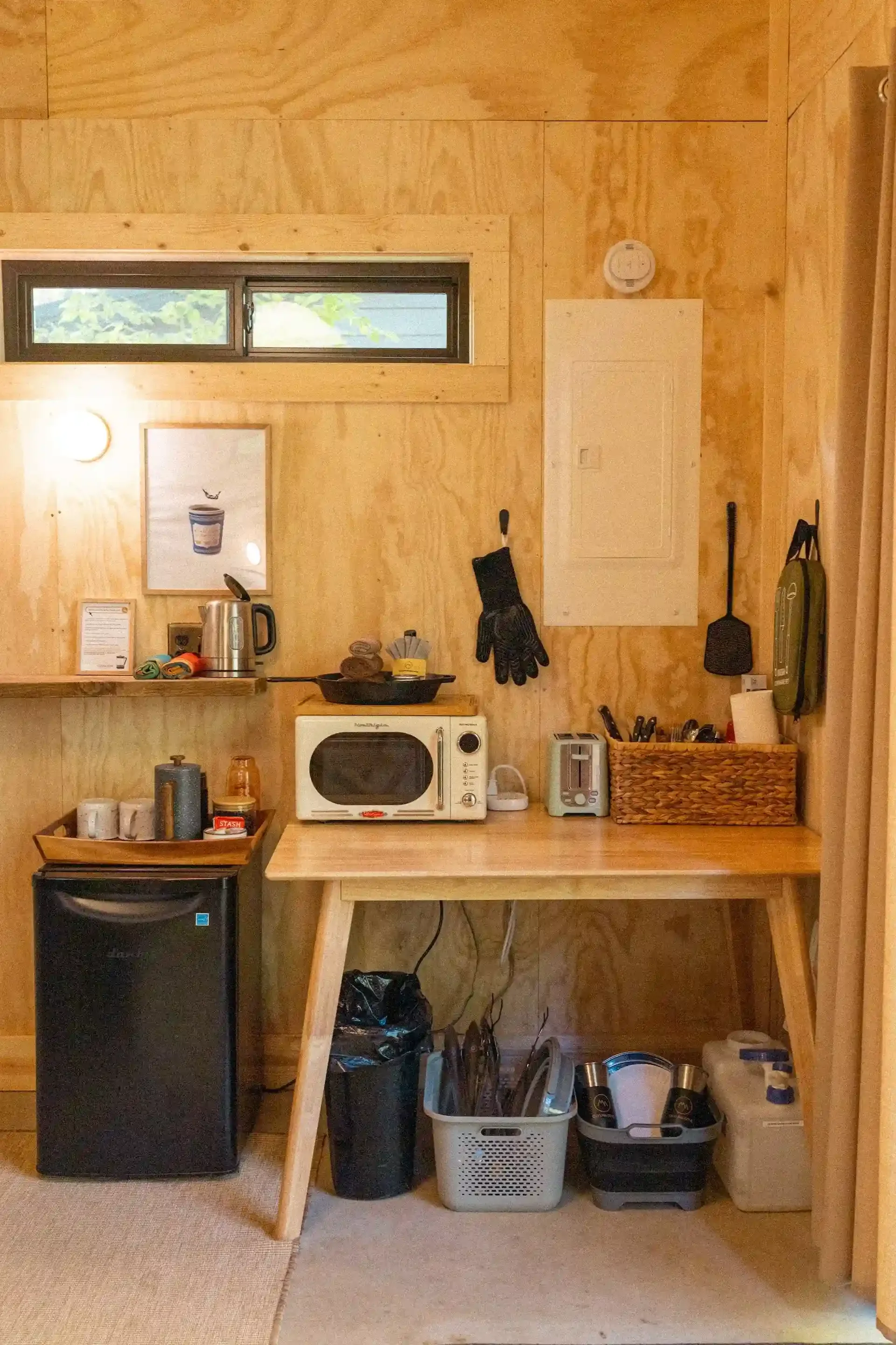 Cozy kitchenette with wooden walls, featuring a small fridge, microwave, kettle, toaster, and kitchen utensils. Baskets and containers hold various items, and a small window and wall art adorn the space. A corkboard and electric panel are mounted on 