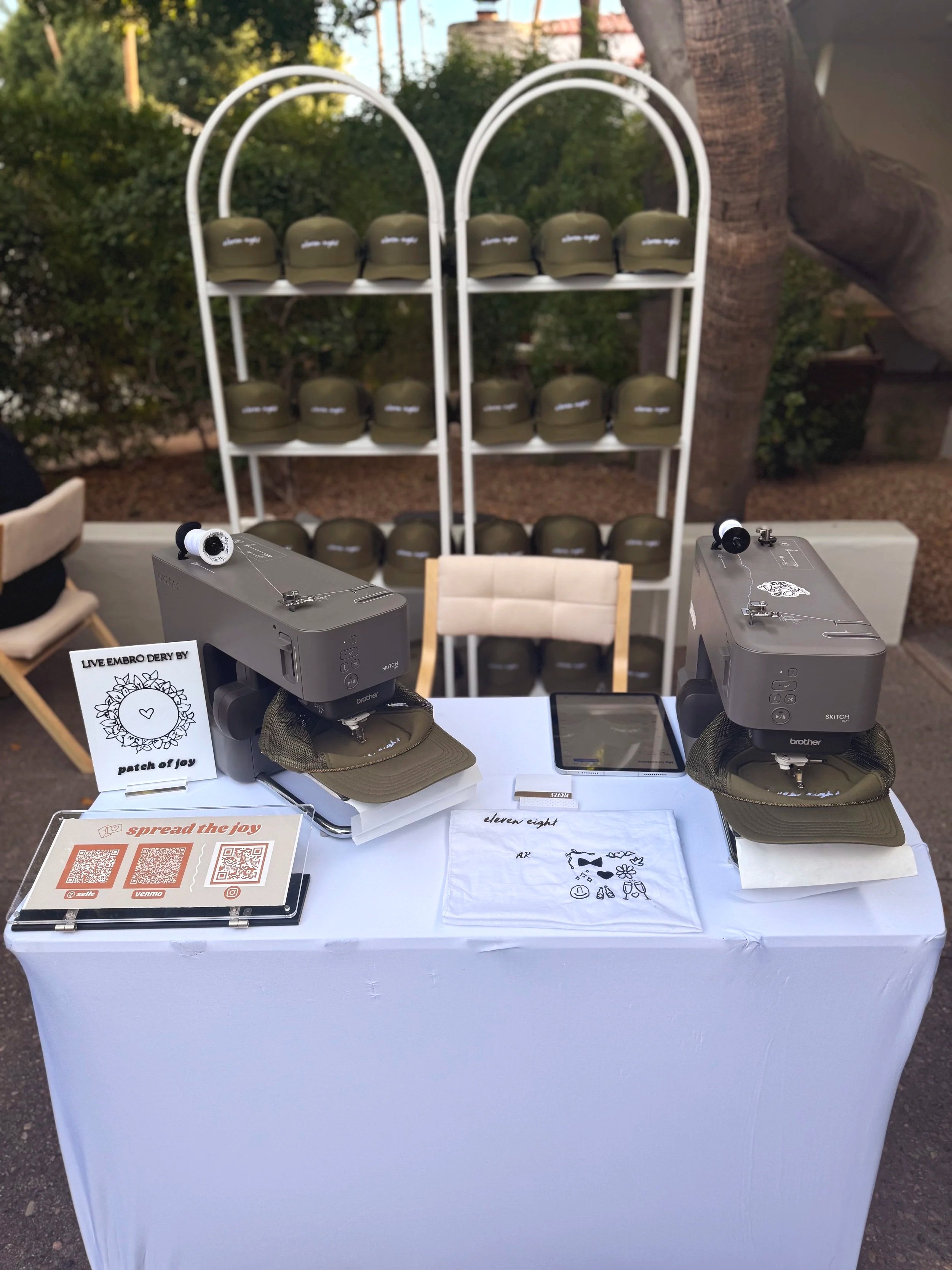Outdoor booth with two sewing machines, each with a cap and spool of thread, a tablet, a cloth with designs, and QR codes on a stand, with a shelf of hats in the background.