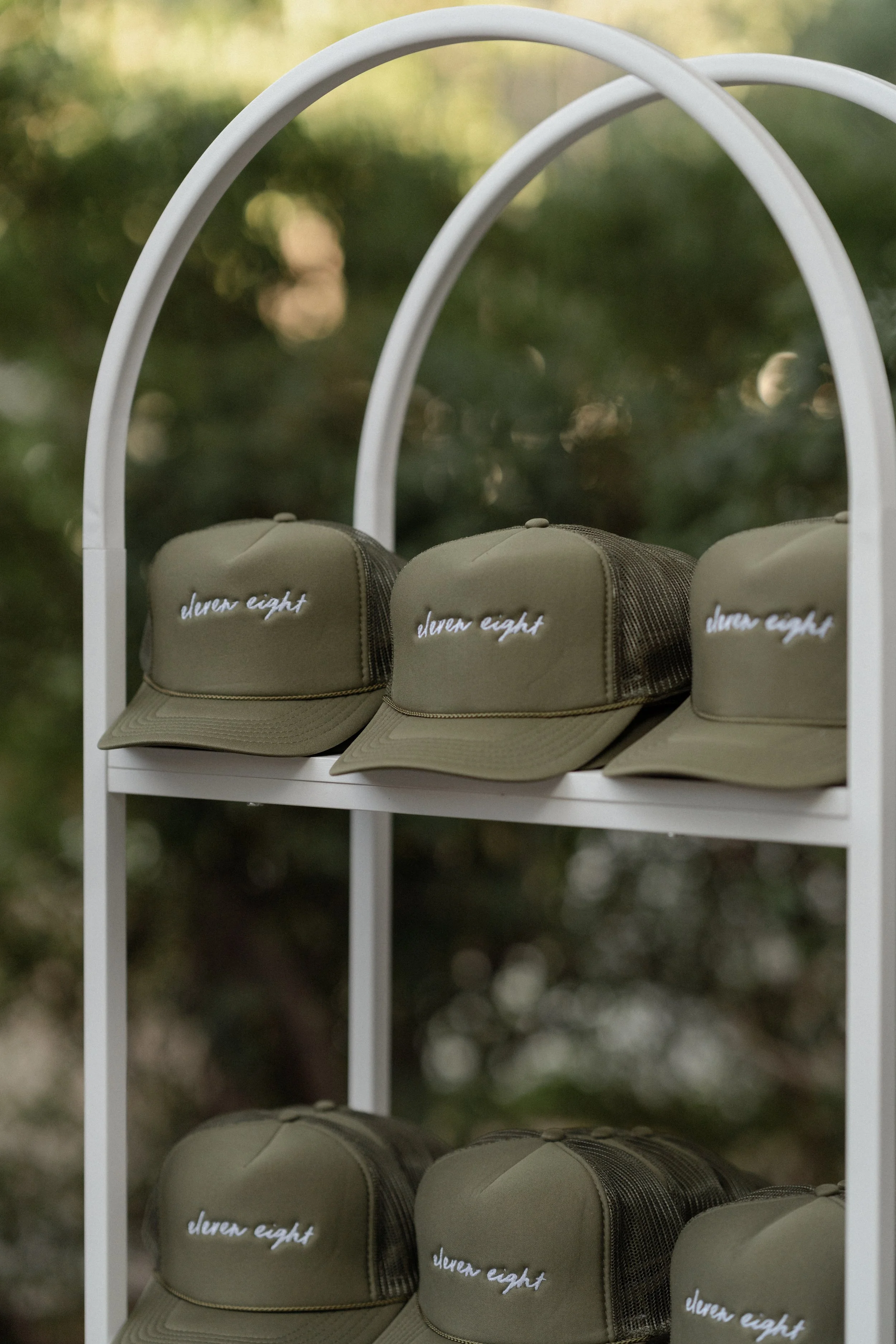 Several olive green caps with white embroidered text 'dleven eight' on a white metal display shelf, outdoors with blurred green leafy background.