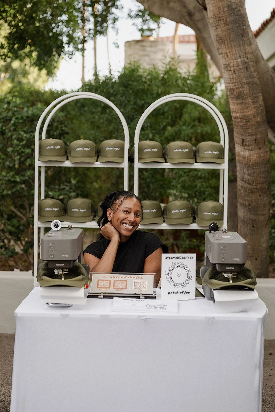 A smiling woman sits at a booth with green hats on display behind her and two machines on the table. The booth is outdoors with trees and a building in the background.