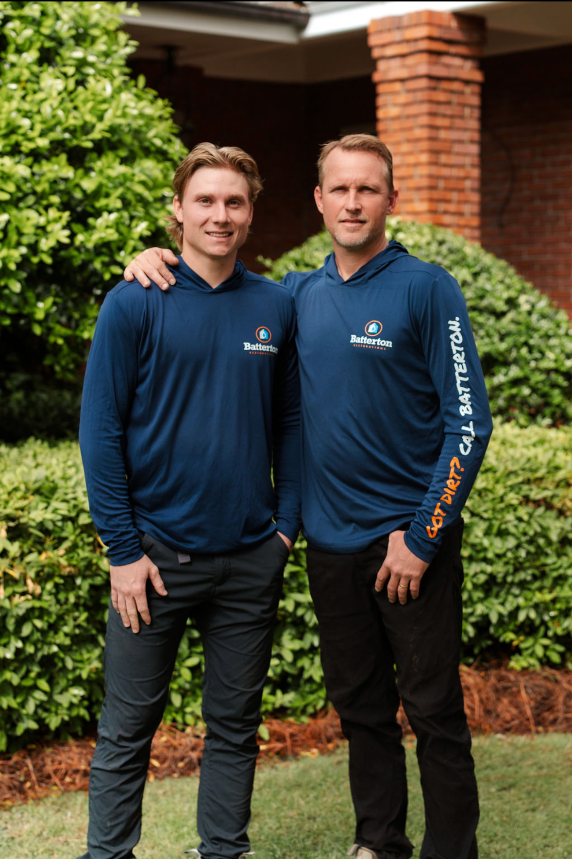 Two men standing outdoors in front of green bushes and a brick building, wearing matching blue long-sleeve shirts with the 'Batterton' logo, posing for a photo.