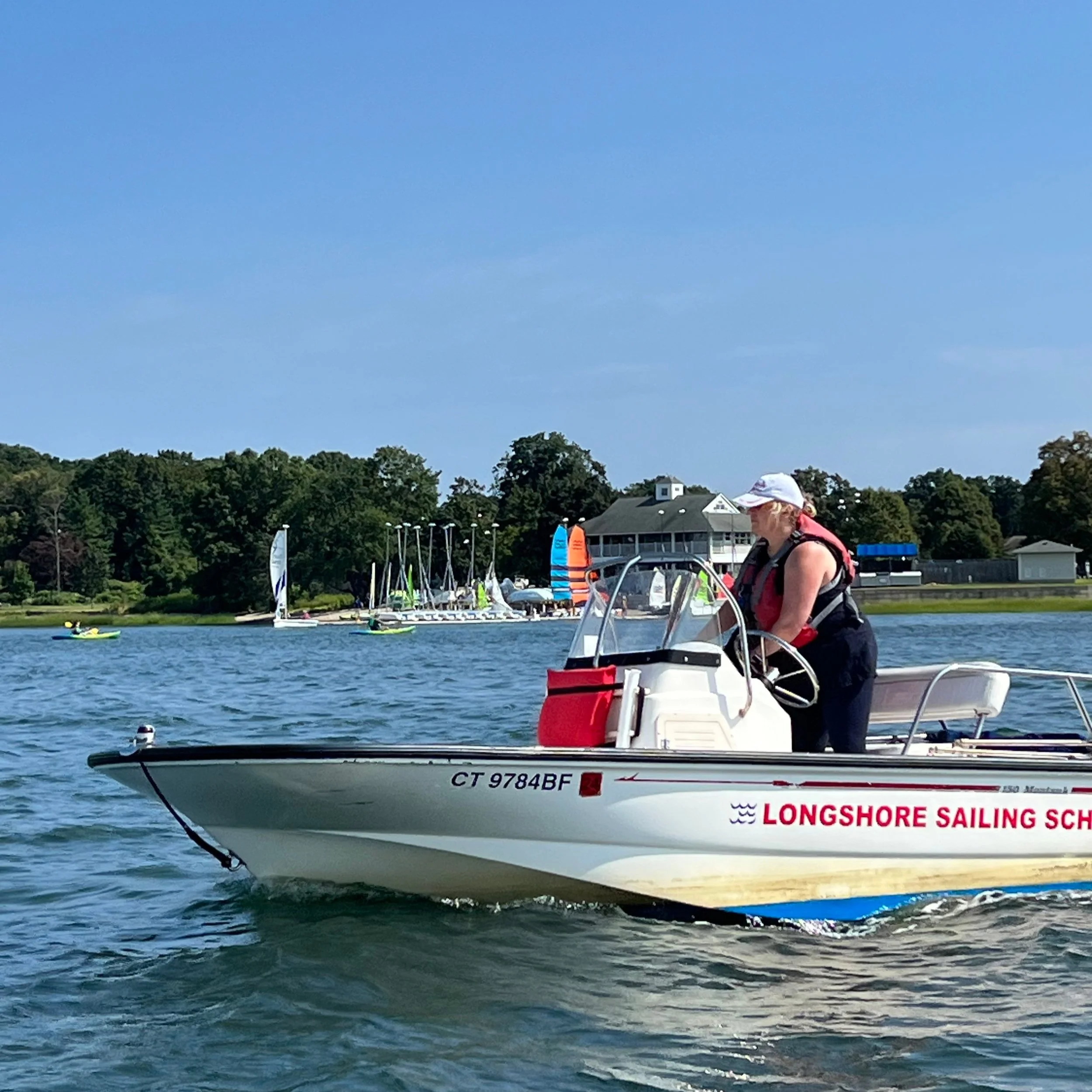 Person operating a motorboat with "Longshore Sailing School" logo, near a shoreline with sailboats and trees in the background.