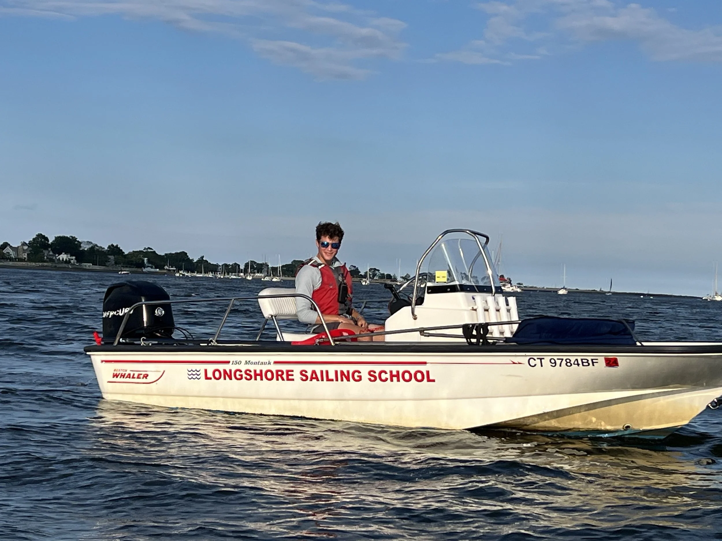Person driving a small motorboat labeled Longshore Sailing School on a body of water.